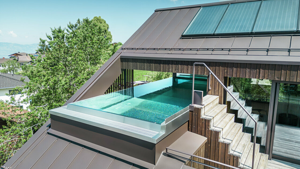 Luftaufnahme eines modernen Hauses mit einem nussbraunen PREFA Prefalz Dach. Eine Dachterrasse mit Pool bietet einen exklusiven Entspannungsbereich mit Blick auf die hügelige, grüne Landschaft.