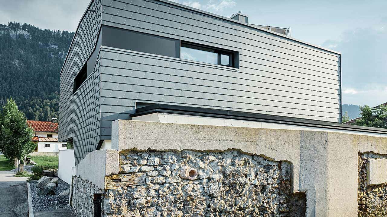 Fassadenverkleidung mit der PREFA Wandschindel aus Aluminium in Hellgrau mit einer Steinmauer vor dem Haus;