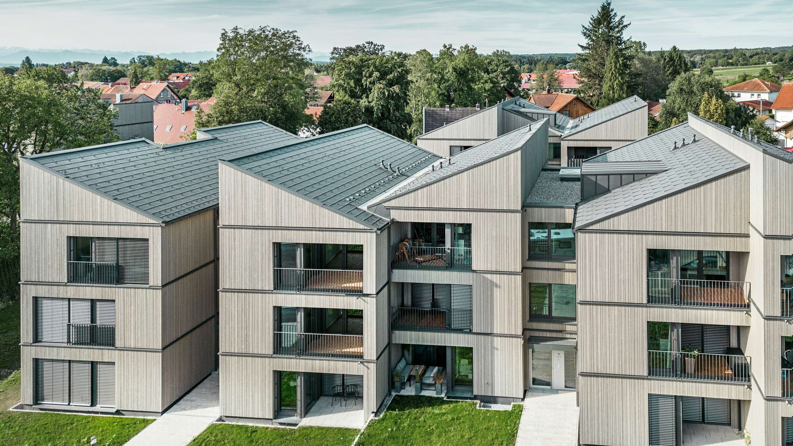 Luftaufnahme des modernen Mehrfamilienhauses am Schmucker-Areal in Utting am Ammersee, mit nachhaltiger Holzhybridbauweise und eleganten Holzfassaden. Die Gebäude sind mit langlebigen PREFA Dachplatten R.16 in P.10 Dunkelgrau ausgestattet. Diese innovative Architektur, geplant von WWA Architekten aus München, zeichnet sich durch klare Linien und harmonische Integration in die grüne Umgebung aus. Die großzügigen Balkone und großen Fenster sorgen für eine helle und freundliche Wohnatmosphäre, während die hochwertigen Materialien für Langlebigkeit und Wetterbeständigkeit sorgen.