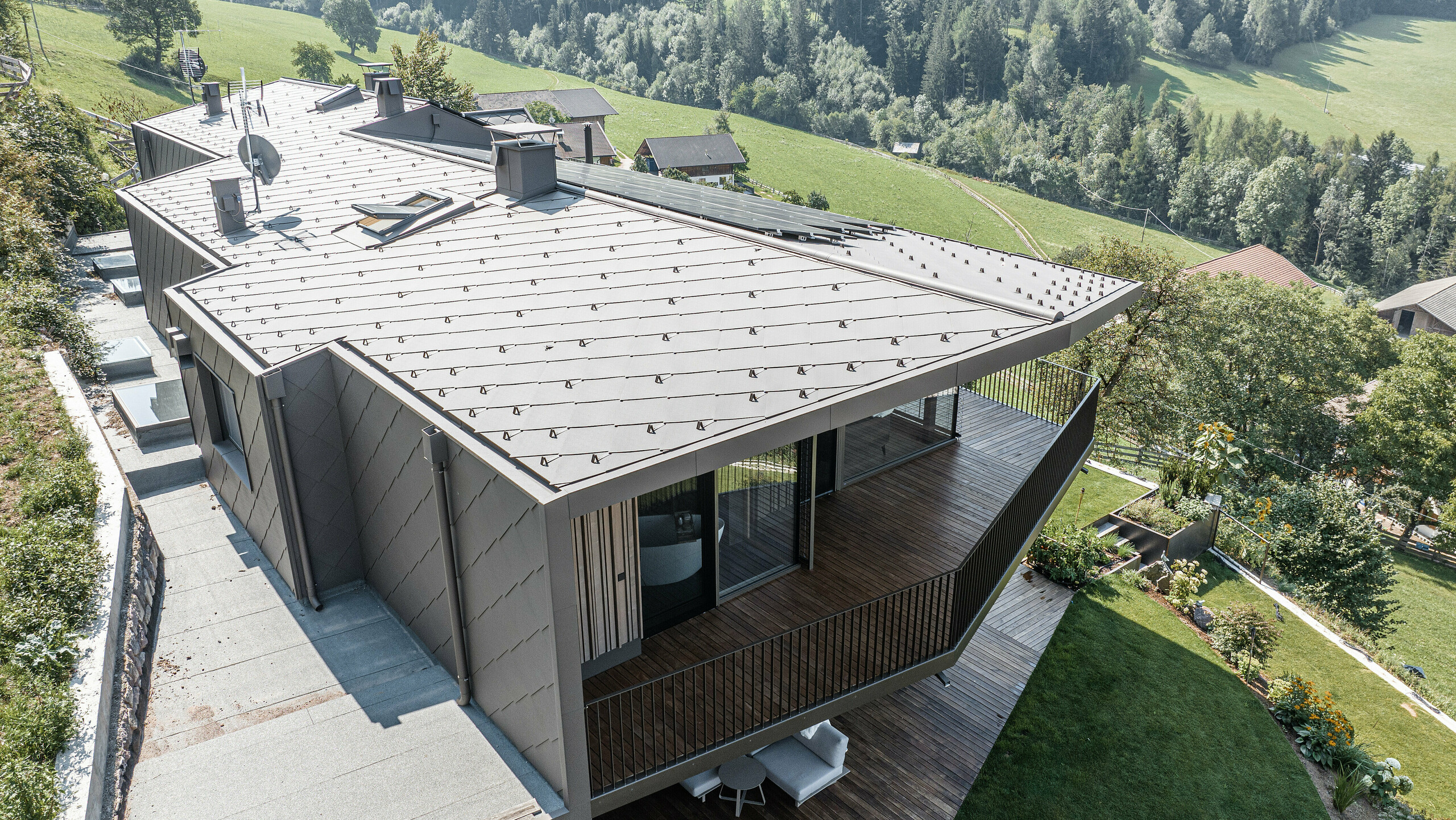 Blick auf eine modernisierte Doppelhaushälfte in Mölten, Südtirol, ausgestattet mit langlebigen PREFA Dach- und Wandrauten 44x44 in P.10 Braun. Die Dach- und Fassadengestaltung kombiniert robustes Aluminium mit Holzelementen, umgeben von einer malerischen Landschaft mit weitem Blick auf bewaldete Hügel. Die Neugestaltung fügt das renovierte Bestandsgebäude und den neu errichteten Standort nahtlos in ein harmonisches Ganzes ein, wobei der Schwerpunkt auf dem Baustoff Holz liegt.