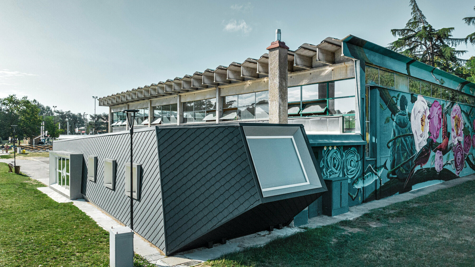 Die Turnhalle Rodriguez und ihr neuer Zubau mit der Rautenschuppenhaut aus PREFA Aluminium von der Seite; der Rasen erstreckt sich davor und zur Seite, im Hintergrund ist ein Teil eines Sportplatzes zu sehen.