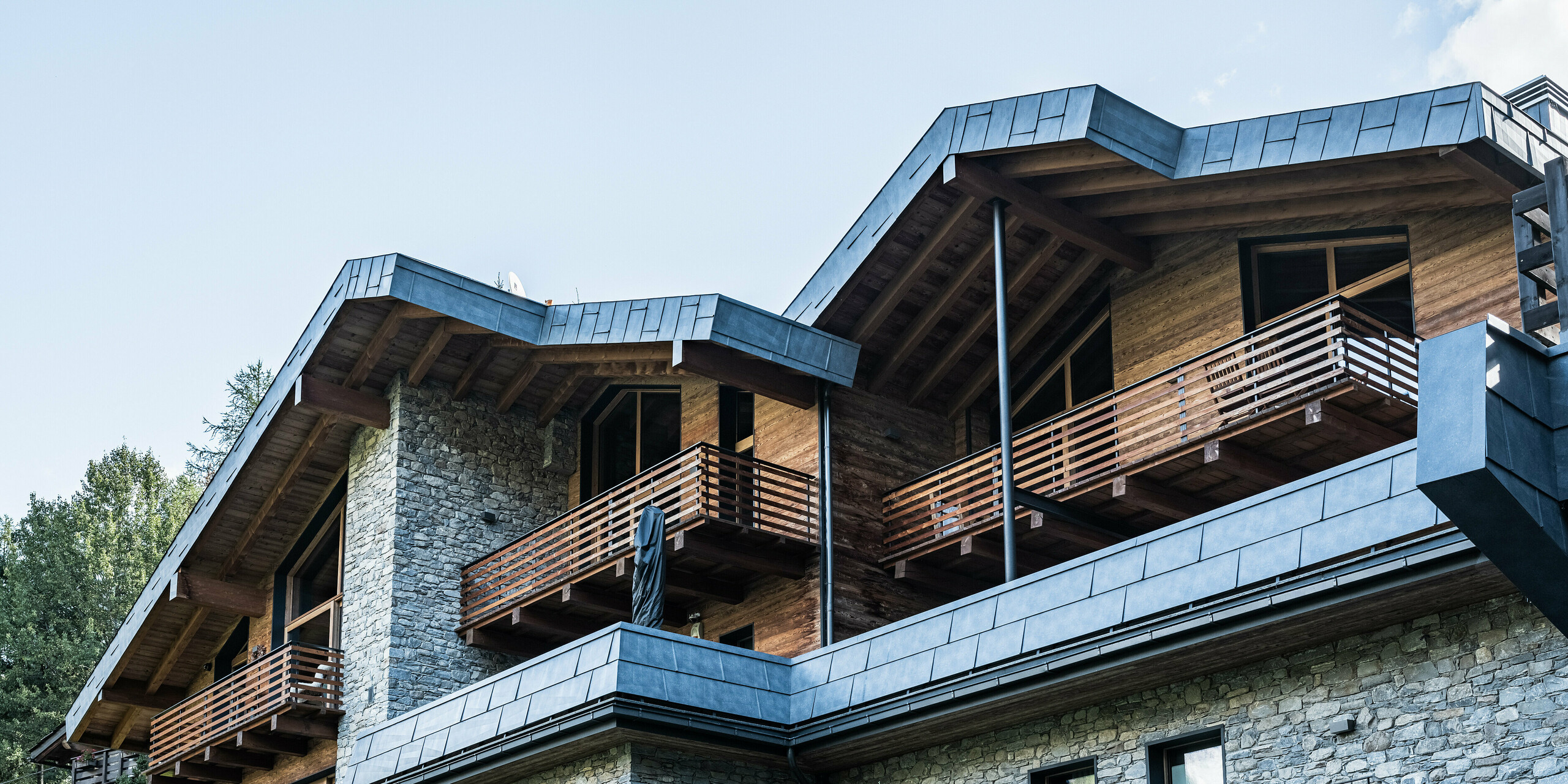 Detailní pohled na moderní bytový dům v Courmayeur s balkony a kombinací stěn z přírodního kamene a dřevěného obkladu. Výrazné střešní a fasádní panely z hliníku PREFA v barvě P.10 břidlicové zdůrazňují vysoce kvalitní architekturu. Konstrukce je rámována okolními borovicemi, které vytvářejí přirozenou a klidnou atmosféru a podtrhují harmonickou souhru moderních a tradičních stavebních prvků.