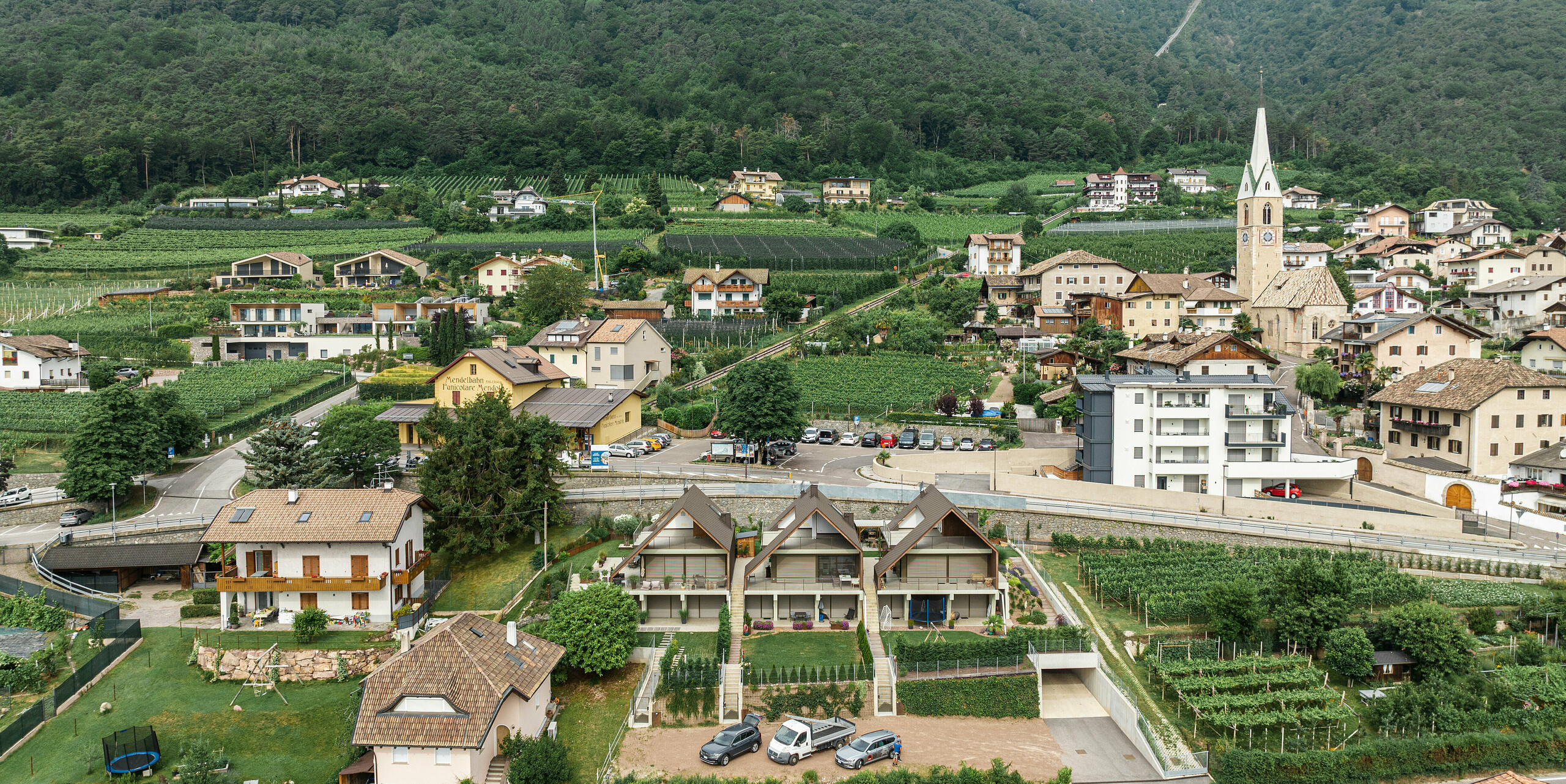 Luftaufnahme eines dicht bebauten Dorfes in einem Tal mit traditionellen und modernen Gebäuden, umgeben von bewirtschafteten Feldern und einem dichten Wald an den Berghängen. Im Zentrum des Dorfes Kaltern in Südtirol steht eine Kirche mit spitzem grünem Turm, die sich von den umliegenden hellen Fassaden und grauen Dächern abhebt. Die Szenerie vermittelt eine harmonische Integration von Architektur und Natur. Im Fokus des Bildes stehen drei Reihenhäuser mit einer Gebäudehülle aus Blech. Die Häuser sind ummantelt von der PREFA Dach- und Wandraute in der Farbe P.10 Braun.