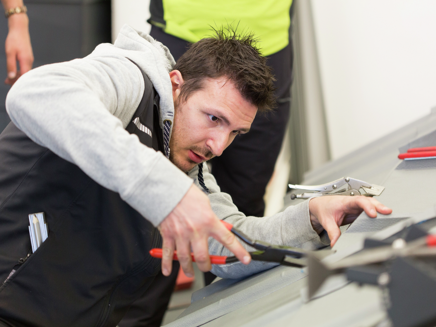 Ein Teilnehmer der PREFA Academy arbeitet konzentriert an einem Aluminiumdachmodell. Er verwendet eine Falzzange. Neben ihm liegen weitere Werkzeuge, die für die Dachinstallation benötigt werden. Die Szene zeigt die praktische Ausbildung und die handwerklichen Fähigkeiten, die in der PREFA Academy vermittelt werden.