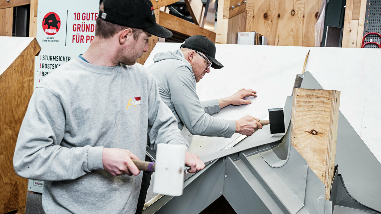 Zwei Teilnehmer des PREFA Academy Kurses arbeiten an einem Modell eines Daches. Sie verwenden Werkzeuge wie Gummihämmer, um Dachelemente aus Aluminium präzise anzubringen. Im Hintergrund ist ein Schild mit den Vorteilen von PREFA-Produkten zu sehen. Die Szene zeigt die praktische Anwendung und den handwerklichen Unterricht in der Akademie.
