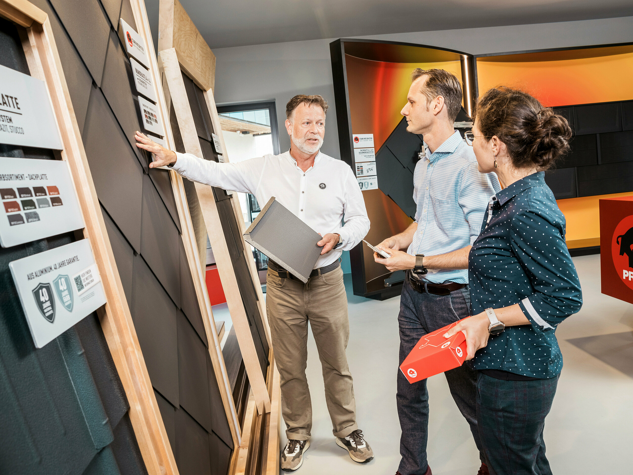 Ein Berater im PREFA Showroom zeigt zwei Kunden die verschiedenen Dachplattenmodelle. Er erklärt die Eigenschaften der Dachplatten in P.10 Anthrazit, während die Kunden aufmerksam zuhören und Prospektmaterialien in der Hand halten.