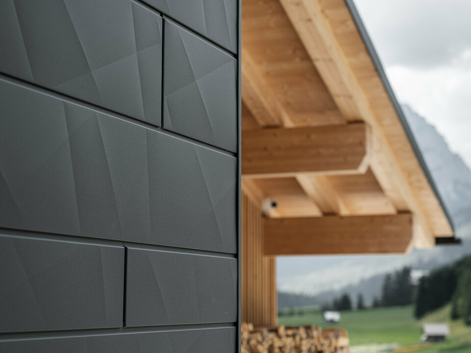 Nahaufnahme der Siding.X Fassadenpaneele in P.10 Schwarz von PREFA an der Casa Hoffe in Sappada, Italien. Das Fassadensystem aus Aluminium ist gekonnt in einem modernen, abstrakten Design angebracht, das einen ansprechenden Kontrast zu den sichtbaren Holzelementen des Dachstuhls bildet. Die innovative Fassadenlösung fügt sich nahtlos in das alpine Ambiente ein und steht für die Verbindung von Funktionalität, Beständigkeit und ästhetischer Baukultur.