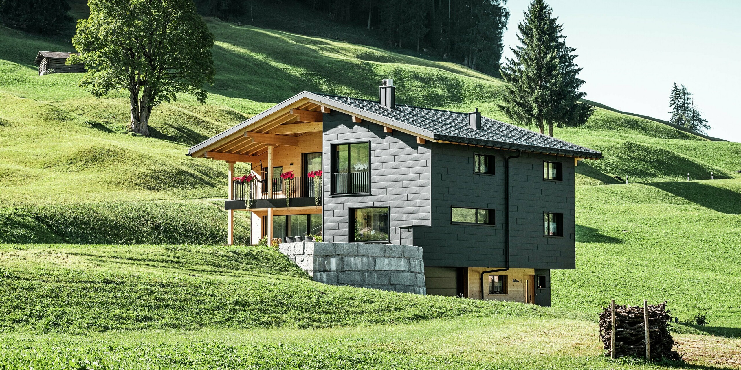 Blick aus der Ferne auf die Terrasse und den Balkon des Hauses, die mit Holz gestaltet sind und die robuste Aluminiumfassade aus PREFA Siding.X in P.10 Schwarz perfekt ergänzen. Die Fassade verleiht dem Gebäude eine moderne Ästhetik und optimale Witterungsbeständigkeit, während die großen Fensterflächen viel Tageslicht ins Innere lassen und die umliegende Natur einrahmen.