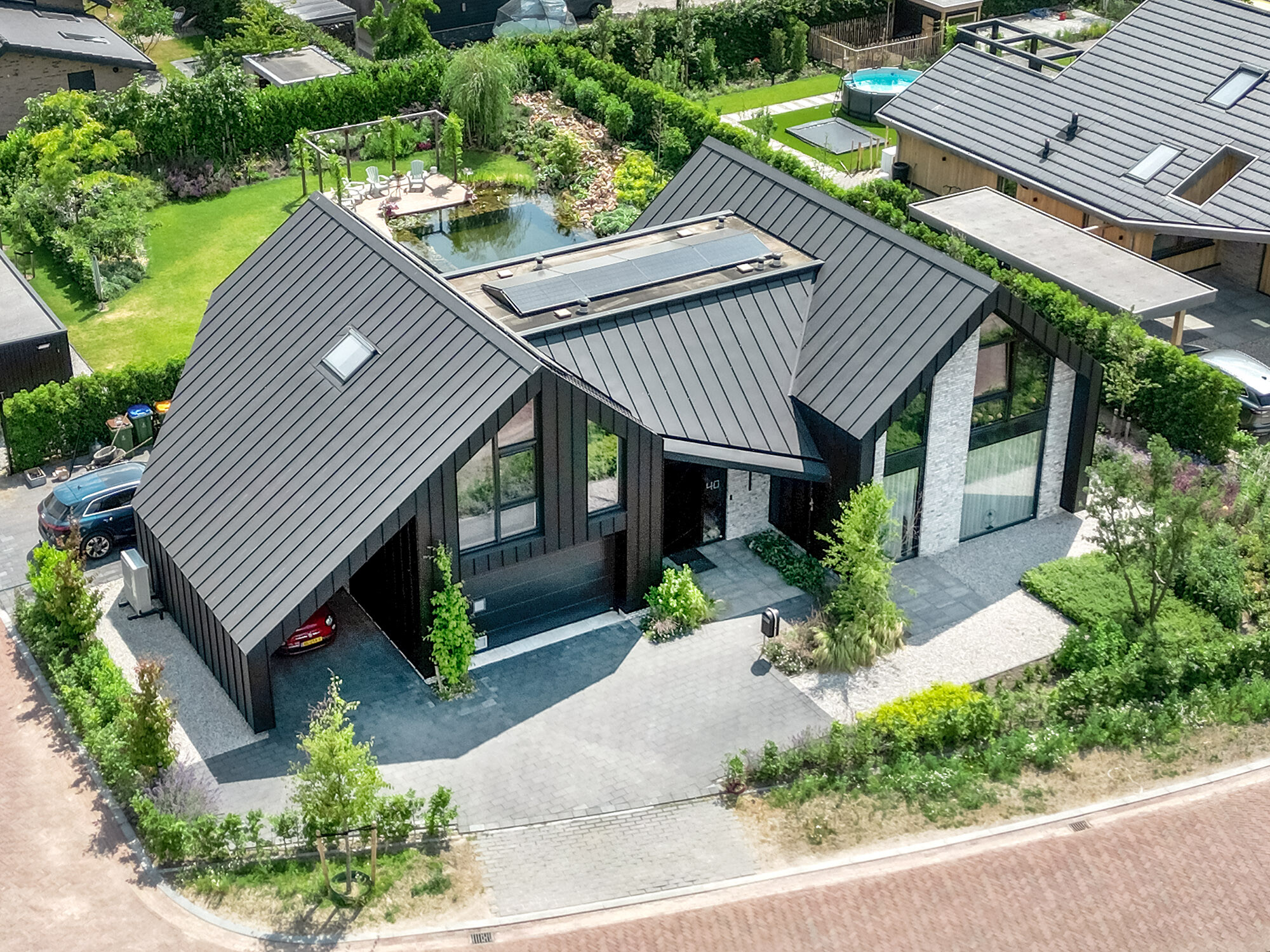 Luftaufnahme eines modernen Einfamilienhauses mit einem steilen, schwarzen Prefalz-Satteldach und integrierten Solarmodulen. Die Architektur kombiniert große Glasfronten und geometrische Formen. Ein Carport ist in das Gebäude integriert. Das Anwesen verfügt über einen gepflegten Garten mit Teich und Terrasse, umgeben von einer ruhigen, vorstädtischen Nachbarschaft.