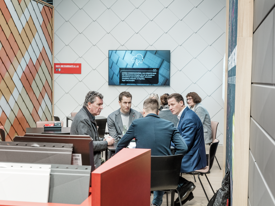 Fachgespräch am PREFA Messestand auf der BAU Messe. Mehrere Personen sitzen an einem Tisch im Beratungsbereich, umgeben von Musterflächen mit Wandrauten in verschiedenen Farbtönen. Ein Bildschirm im Hintergrund zeigt den PREFA Messefilm.