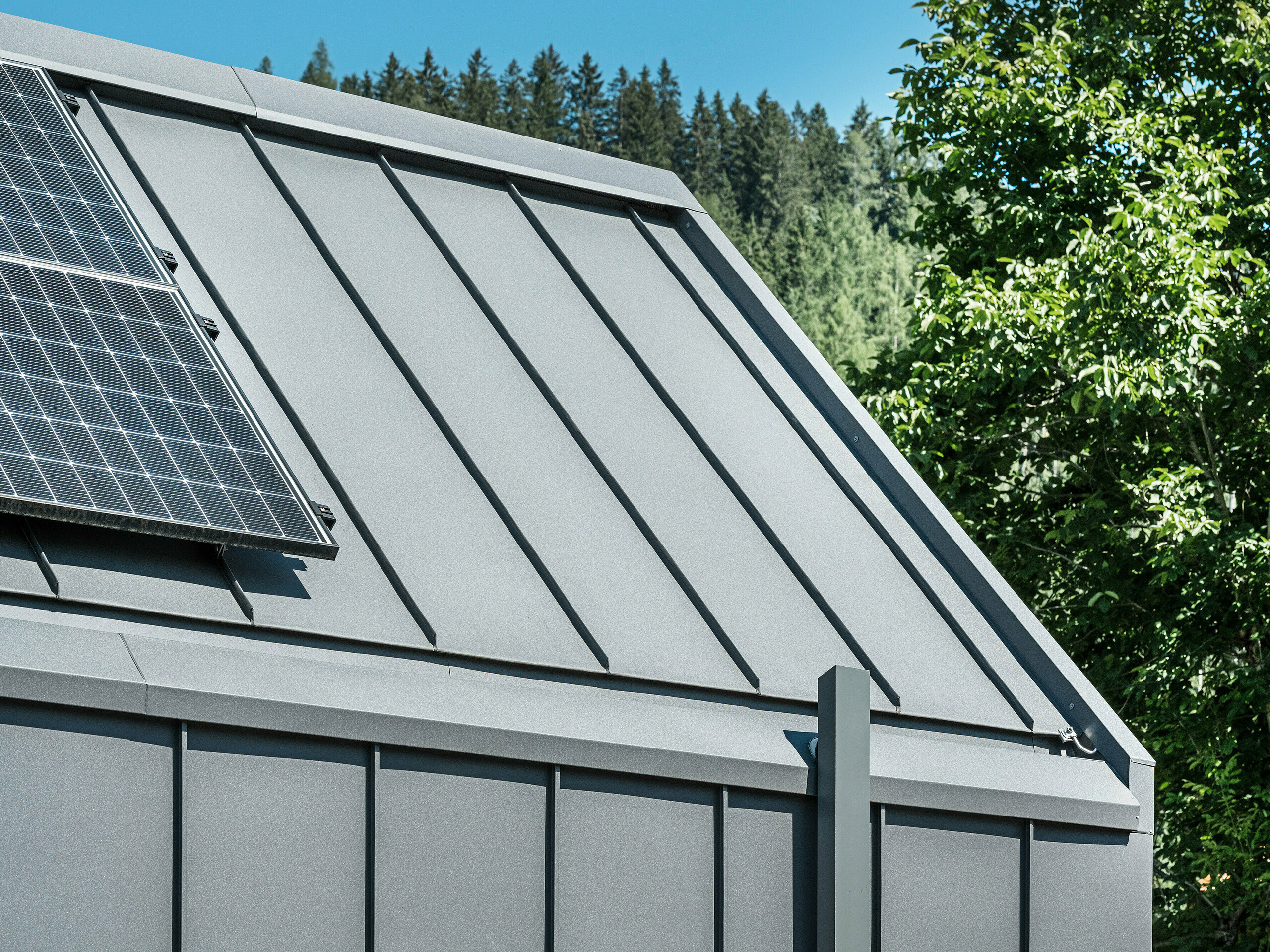 Nahaufnahme von Dach und Fassade eines modernen Einfamilienhauses in Pogier, Österreich, mit einer nachhaltigen und langlebigen Aluminiumhülle von PREFA. Dach und Fassade sind mit PREFALZ in P.10 Dunkelgrau eingedeckt, was dem Haus ein elegantes und zeitloses Aussehen verleiht. Darüber hinaus ist das Haus mit einer hochwertigen Dachentwässerung ausgestattet, die Funktionalität und Ästhetik in der Architektur perfekt vereint. Die Dachentwässerung erfolgt über das PREFA Quadratrohr - ebenfalls in Dunkelgrau gehalten sind, und die am linken Bildrand sichtbare Photovoltaikanlage am Dach unterstreicht das nachhaltige Konzept des Gebäudes. Im Hintergrund sind grüne Bäume und blauer Himmel zu sehen.