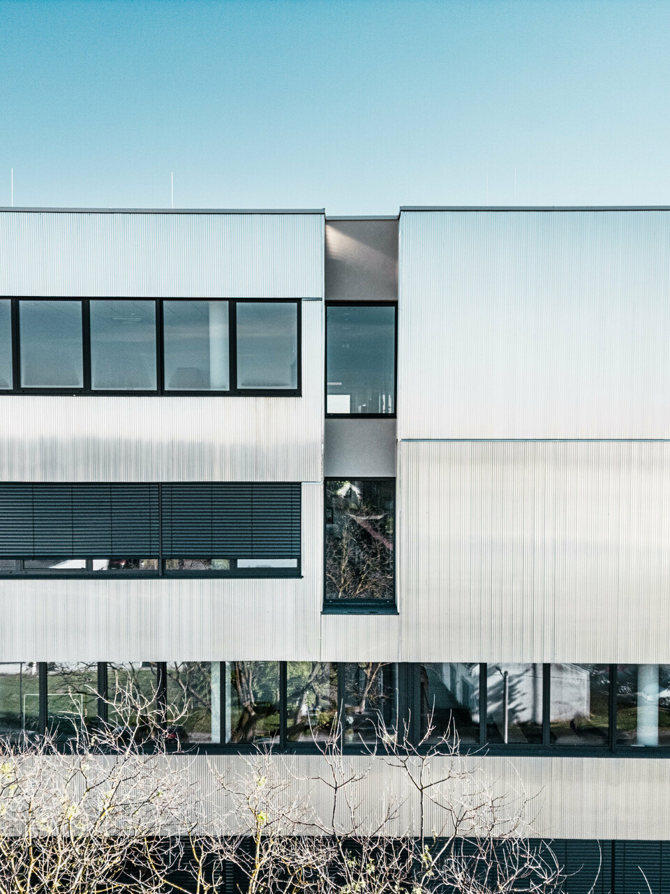 Eine moderne mehrstöckige Musikschule in Bjevolar mit einer PREFA Fassade im Zackenprofil. Die Fassade ist teilweise in Schwarz und aluminiumblank gehalten und reflektiert das Sonnenlicht, während große Glasfenster für einen modernen Look sorgen. Im Hintergrund sind Wohngebäude und Bäume sichtbar.