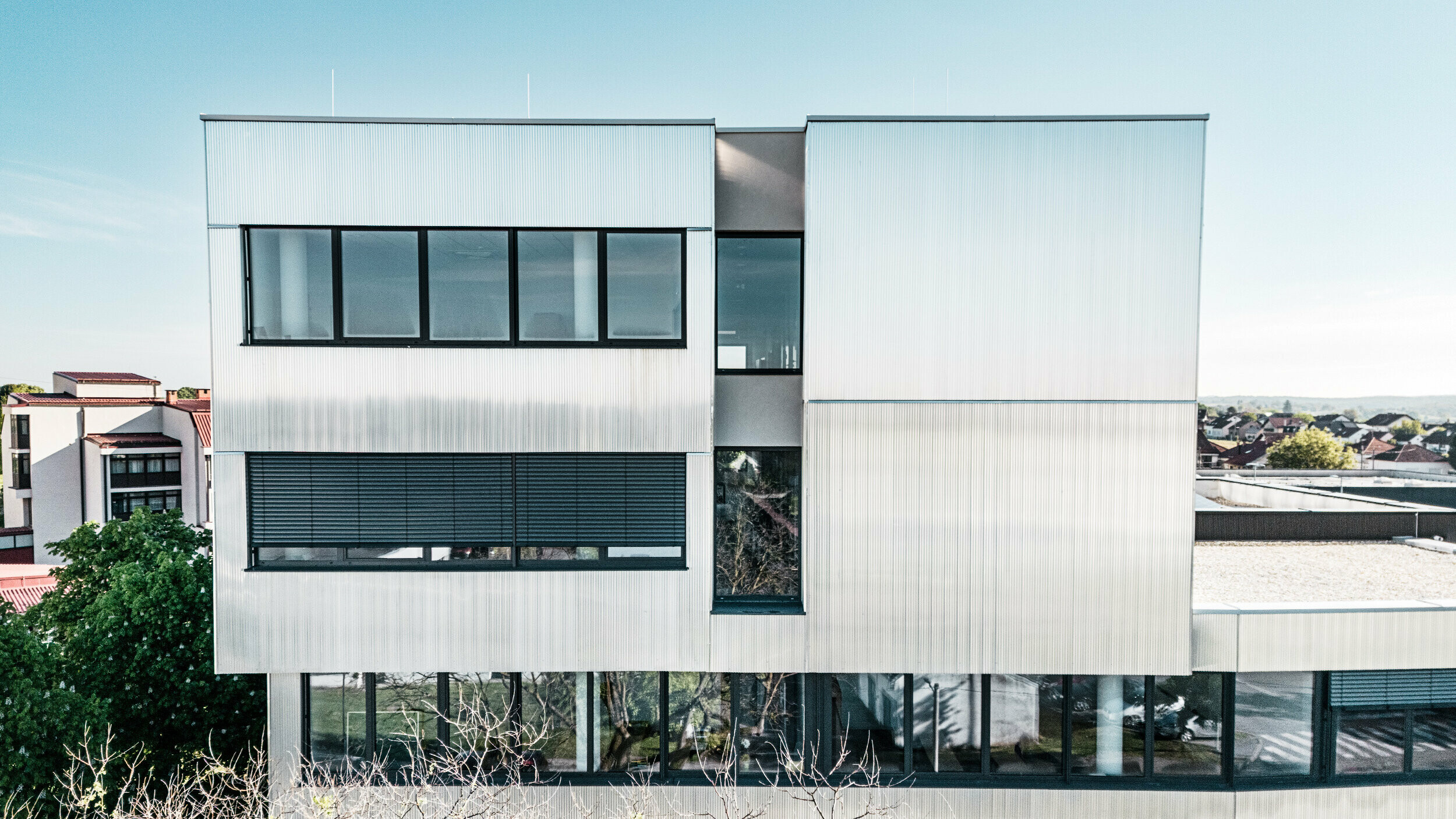 Eine moderne mehrstöckige Musikschule in Bjevolar mit einer PREFA Fassade im Zackenprofil. Die Fassade ist teilweise in Schwarz und aluminiumblank gehalten und reflektiert das Sonnenlicht, während große Glasfenster für einen modernen Look sorgen. Im Hintergrund sind Wohngebäude und Bäume sichtbar.