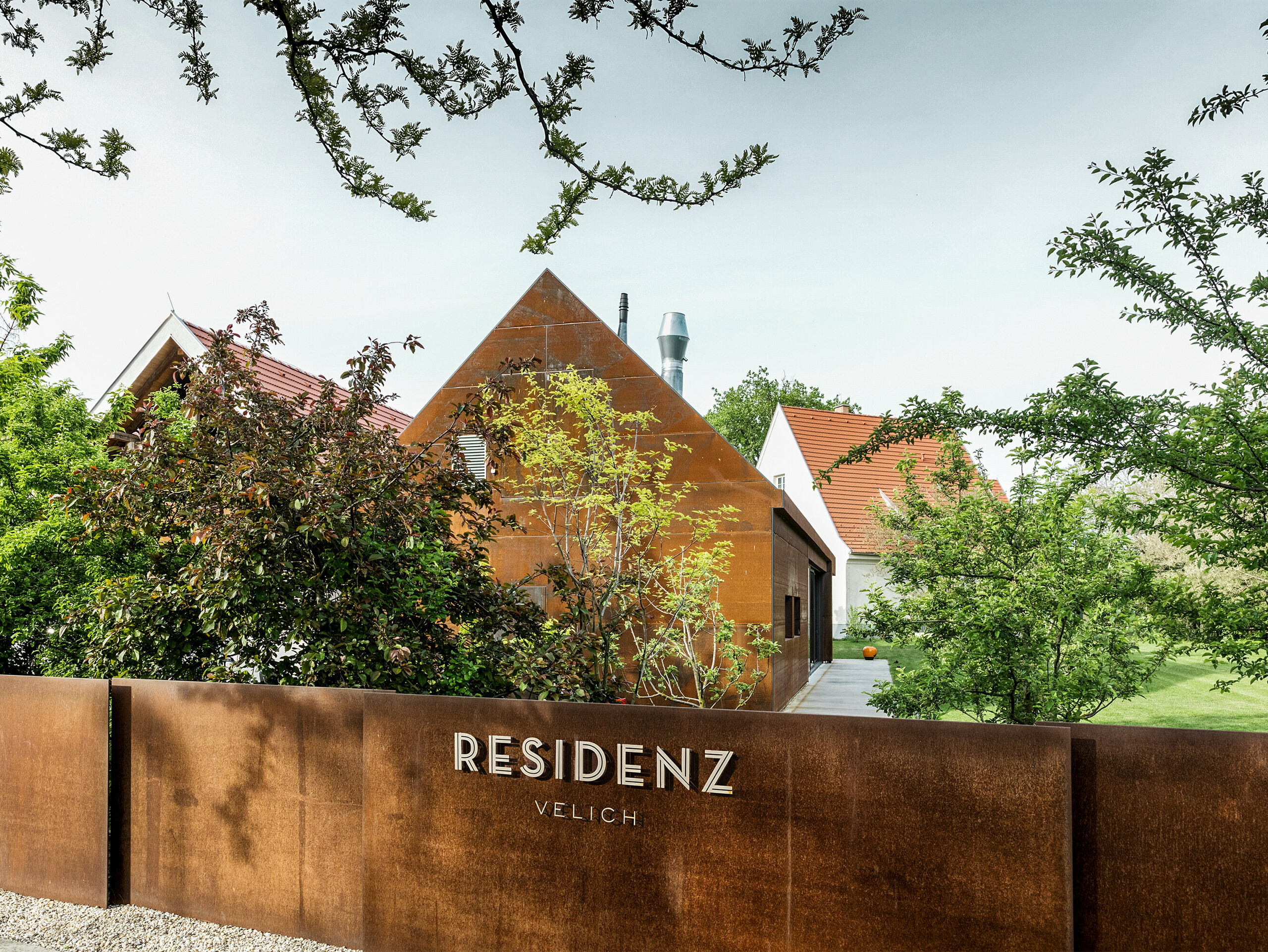 Wohnhaus Velich im Seewinkel, Burgenland: Blick auf die moderne Gartenanlage mit schönen Pflanzen. Das Wohnhaus dient als Veranstaltungsort für den Fachdialog PREFARENZEN 2025, bei dem Architekten und PREFA-Experten die interessantesten Architekturobjekte auswählen. Im Vordergrund ist eine stilvolle, rostfarbene Metallwand mit der Aufschrift „RESIDENCE VELICH“ zu sehen. Die Architektur des Gebäudes verbindet traditionelle und moderne Elemente und fügt sich harmonisch in die natürliche Umgebung ein.