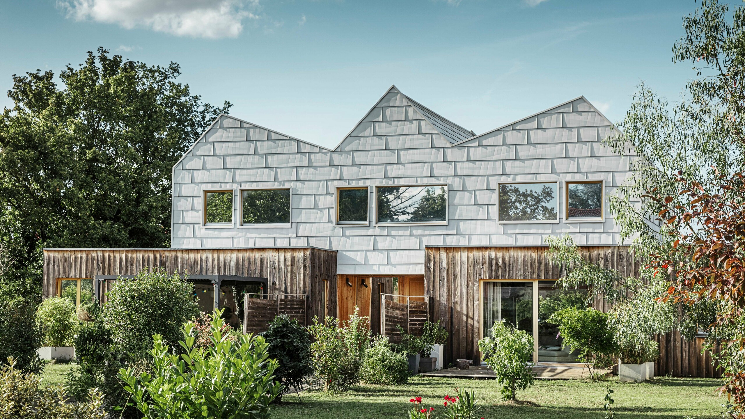 Modernes Doppelhaus mit Satteldach und PREFA-Fassade. Die Fassade besteht aus FX.12 Aluminiumpaneelen, die eine strukturierte, moderne Optik bieten. Das Haus ist von einem gepflegten Garten umgeben, und die Fensterrahmen harmonieren perfekt mit der Gesamtarchitektur.