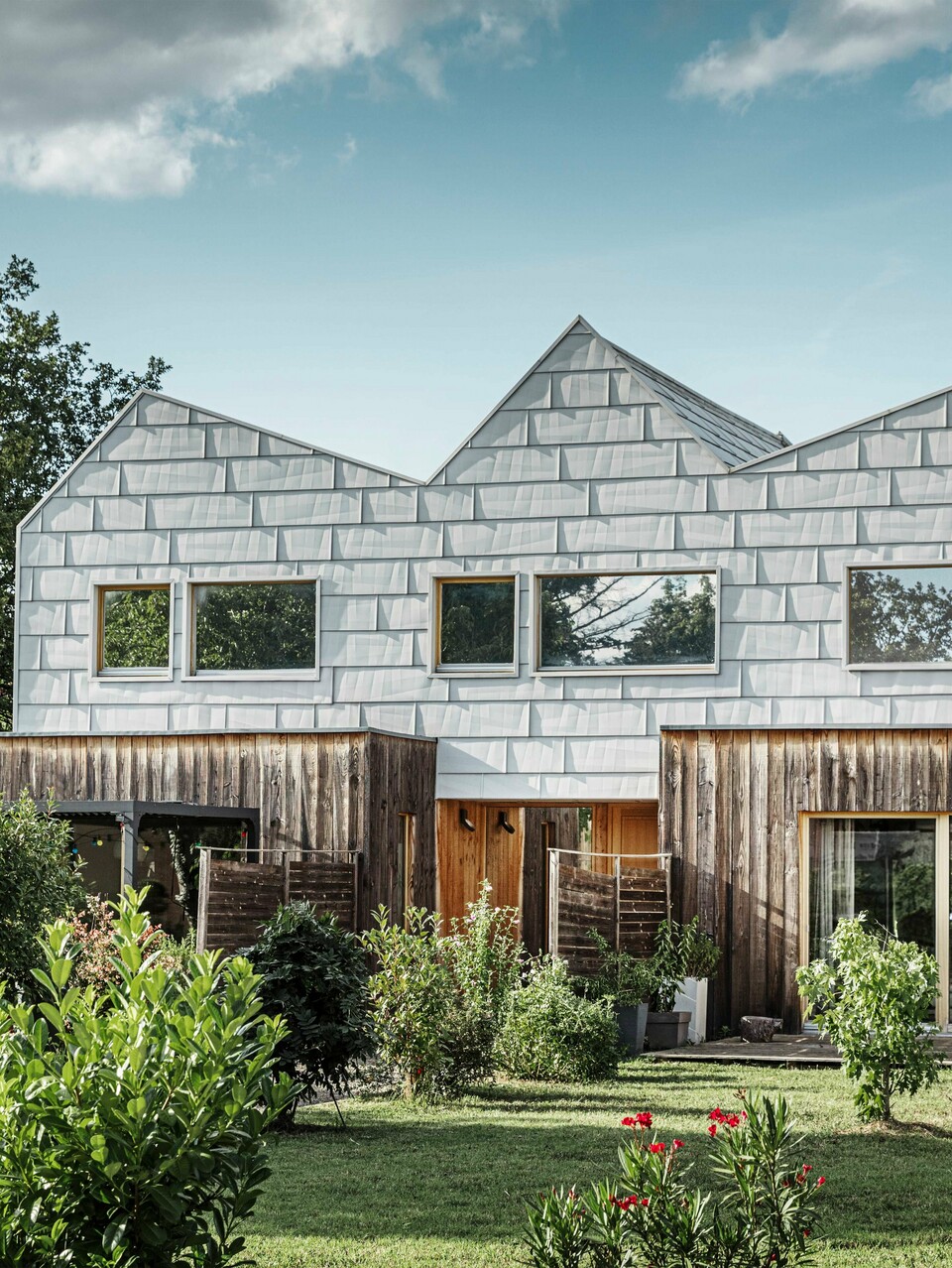 Modernes Doppelhaus mit Satteldach und PREFA-Fassade. Die Fassade besteht aus FX.12 Aluminiumpaneelen, die eine strukturierte, moderne Optik bieten. Das Haus ist von einem gepflegten Garten umgeben, und die Fensterrahmen harmonieren perfekt mit der Gesamtarchitektur.