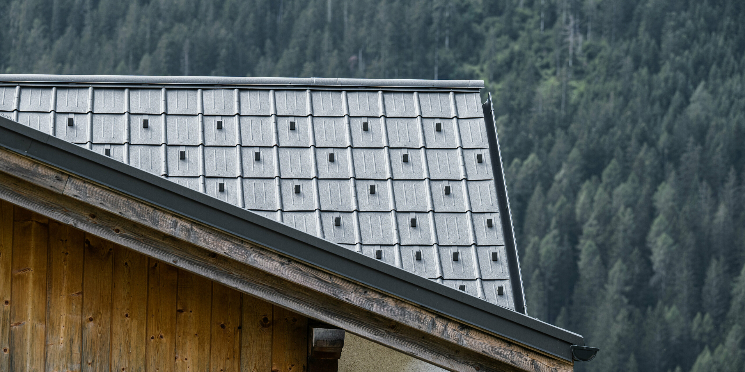 Nahaufnahme eines Aluminiumdachs mit hellgrauen PREFA-Ziegeln auf einer Berghütte in Auronzo di Cadore, Italien, umgeben von dichten Wäldern. Die präzise Verlegung und das einfache Design der Dachplatten unterstreichen die ländliche Architektur und bieten gleichzeitig modernen Schutz und Ästhetik im Einklang mit der natürlichen Bergumgebung.