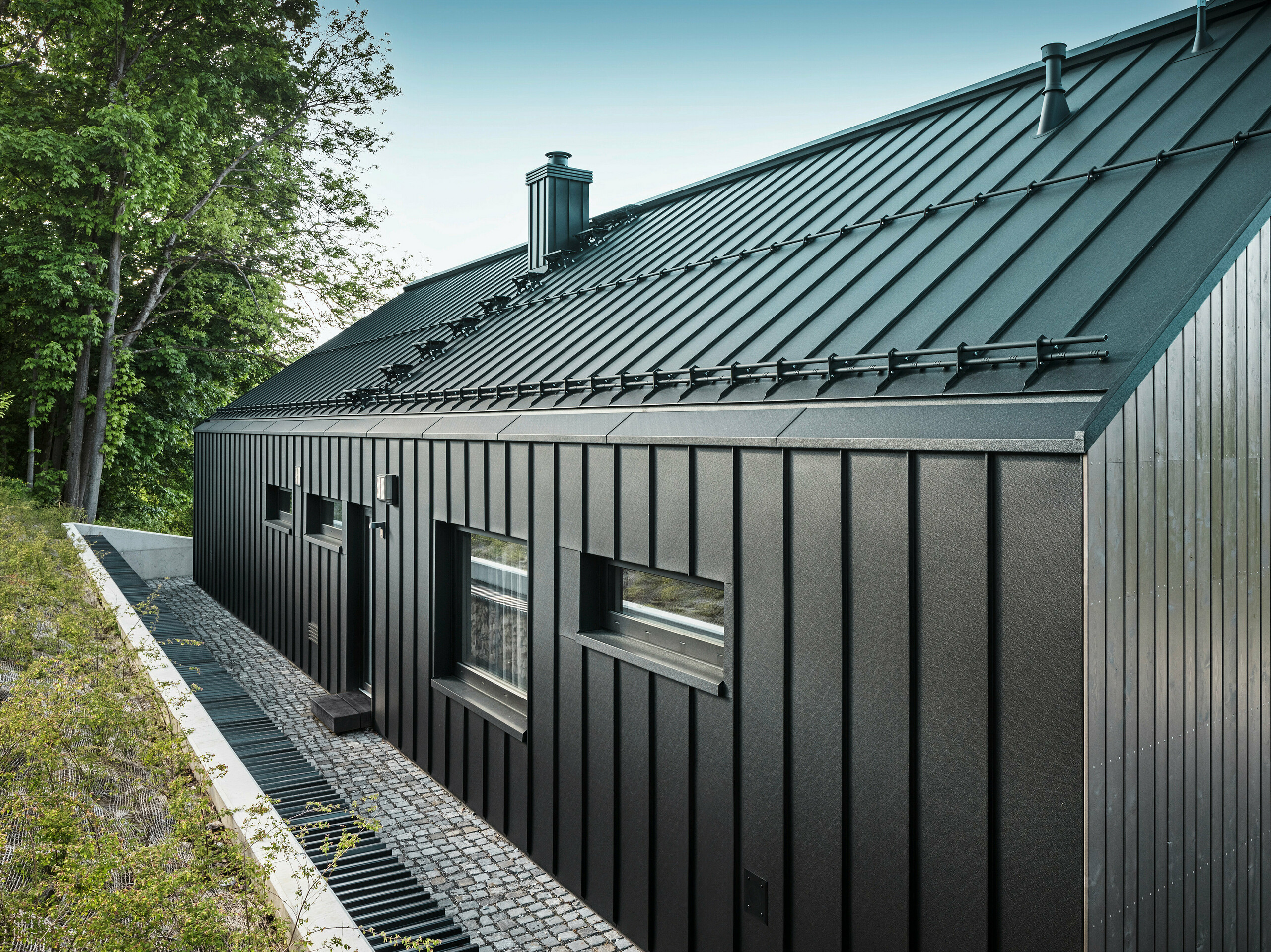 Nahaufnahme der Rückseite eines modernen Einfamilienhauses in Kuncice, CZ. Das dunkle Holzmassivhaus wurde am Dach und der Gebäuderückseite großflächig mit PREFALZ in P.10 Schwarz verkleidet. Auf der Rückseite sind drei horizontale sowie ein quadratisches Fenster angebracht worden. Zwischen den vier Fenstern ist eine Tür mit einer kleinen Stufe zu sehen.