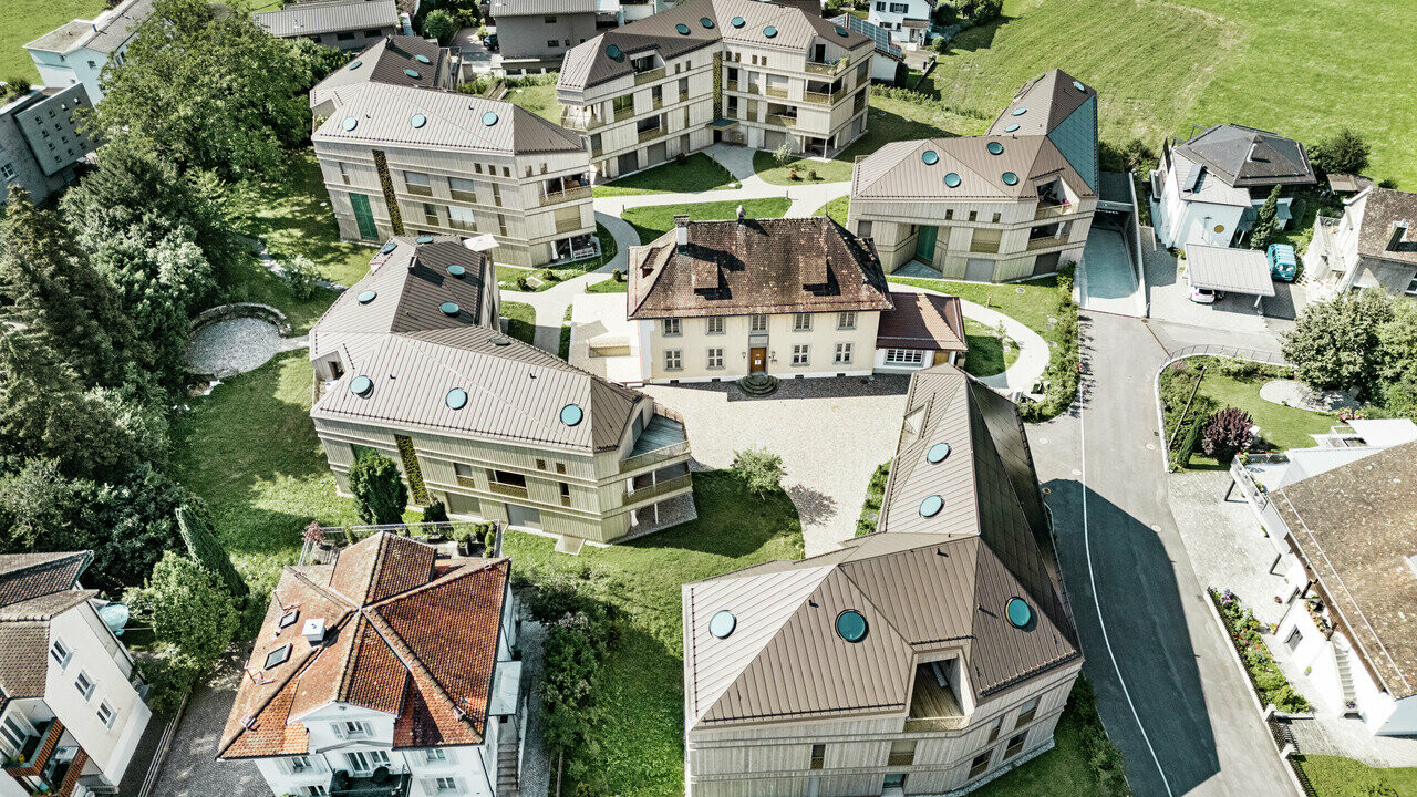 Luftaufnahme der modernen Wohnanlage in Liebwylen, Schweiz. Die Gebäude sind mit PREFALZ Aluminiumdächern in Braun gestaltet und verfügen über runde Dachfenster. Im Zentrum steht die historische Villa, umgeben von harmonisch angeordneten Neubauten, eingebettet in eine grüne Landschaft.