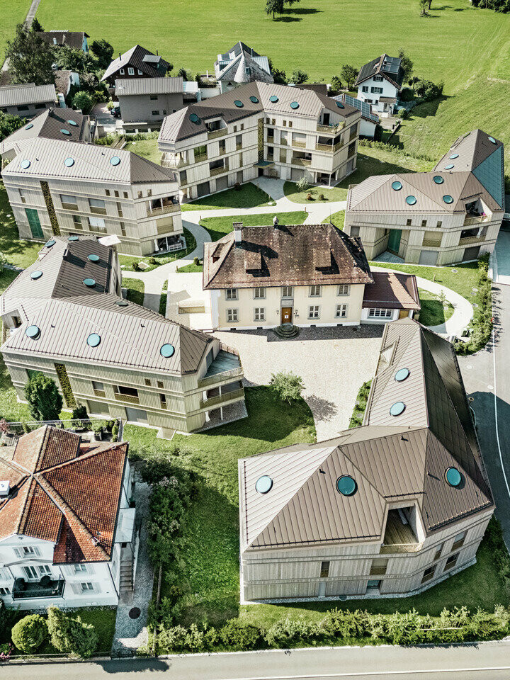 Luftaufnahme der modernen Wohnanlage in Liebwylen, Schweiz. Die Gebäude sind mit PREFALZ Aluminiumdächern in Braun gestaltet und verfügen über runde Dachfenster. Im Zentrum steht die historische Villa, umgeben von harmonisch angeordneten Neubauten, eingebettet in eine grüne Landschaft.