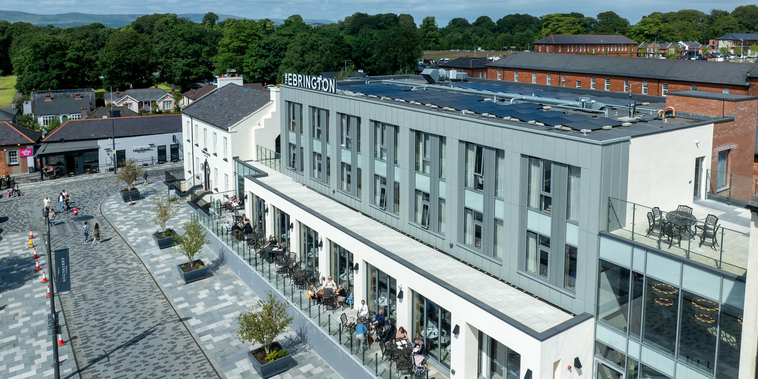 Das Ebrington Hotel im Zentrum von Londonderry, Nordirland, überzeugt seine moderne Fassade aus Aluminium. Das Obergeschoss ist mit PREFALZ in den Farben P.10 Hellgrau und P.10 Zinkgrau verkleidet, was dem Gebäude eine elegante und robuste Optik verleiht. Im Vordergrund ist die belebte Terrasse des Hotels zu sehen, wo Gäste entspannen und die Umgebung genießen können. Die hochwertige Blechverkleidung trägt zur ästhetischen und langlebigen Erscheinung des Hotels bei.