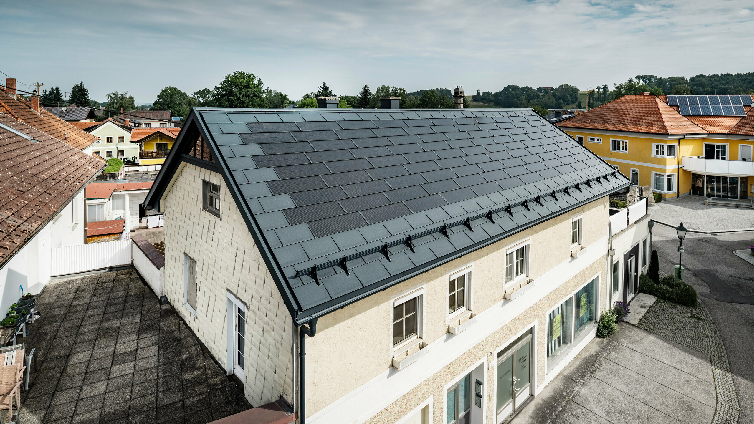 Blick von oben auf ein Einfamilienhaus in Mettmach, Österreich. Das ältere Gebäude wurde mit anthrazitfarbenen PREFA Solardachplatten saniert. Die Schneefangsysteme und die klare Linienführung der Solardachplatten unterstreichen die saubere Verarbeitung der PREFA Aluminiumprodukte. Im Hintergrund kontrastiert das orangefarbene Ziegeldach eines Nachbargebäudes. Auf dem Dach des benachbarten Einfamilienhauses sind Photovoltaikmodule in einer klassischen Aufdach-Lösung installiert, was die Vielfalt der energieeffizienten Dachgestaltung zeigt.