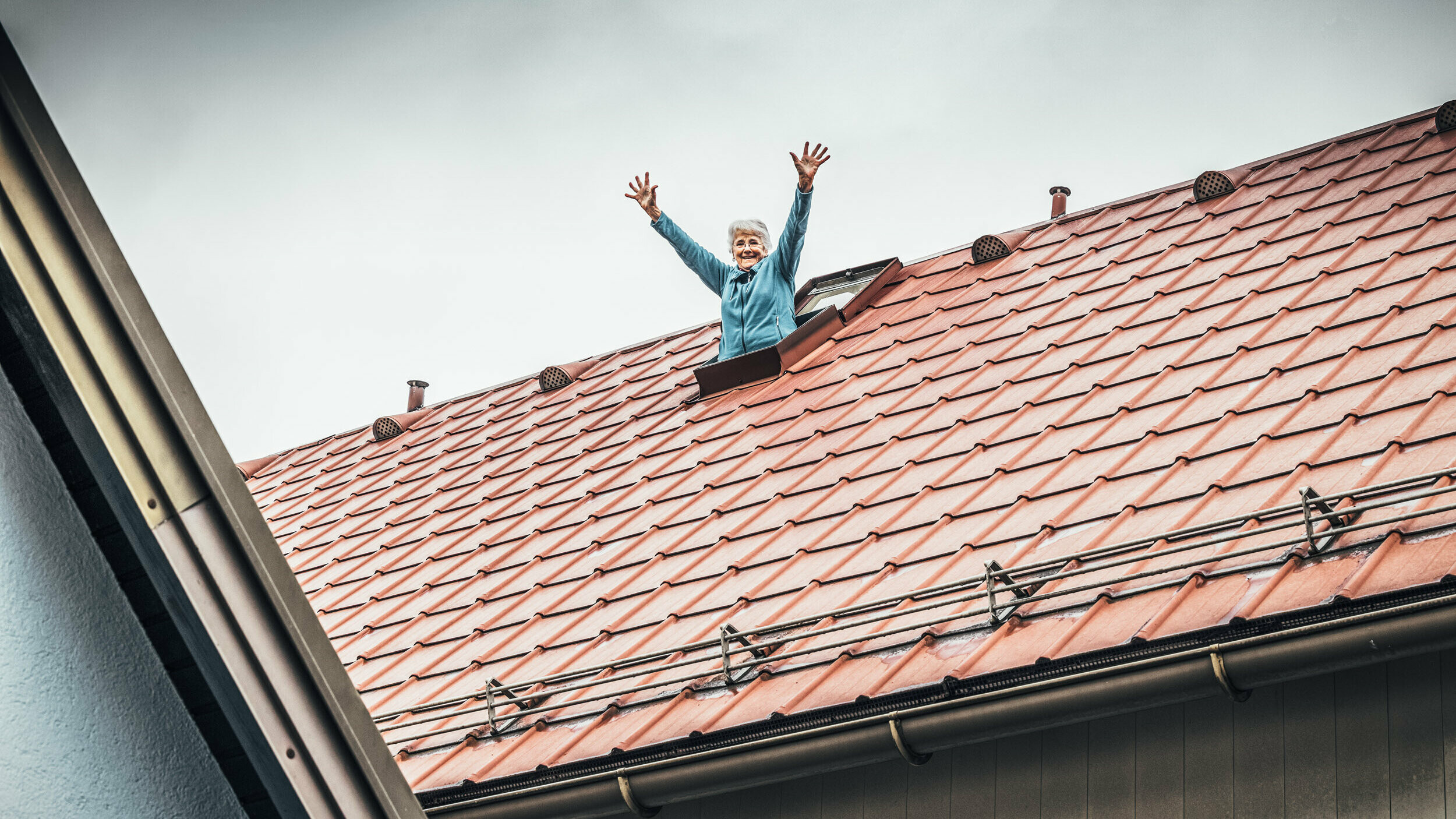 Anna streckt den Oberkörper mit ausgestreckten Armen aus einem Dachflächenfenster und lächelt. Das Dach wurde mit der PREFA Dachplatte in Ziegelrot eingedeckt.