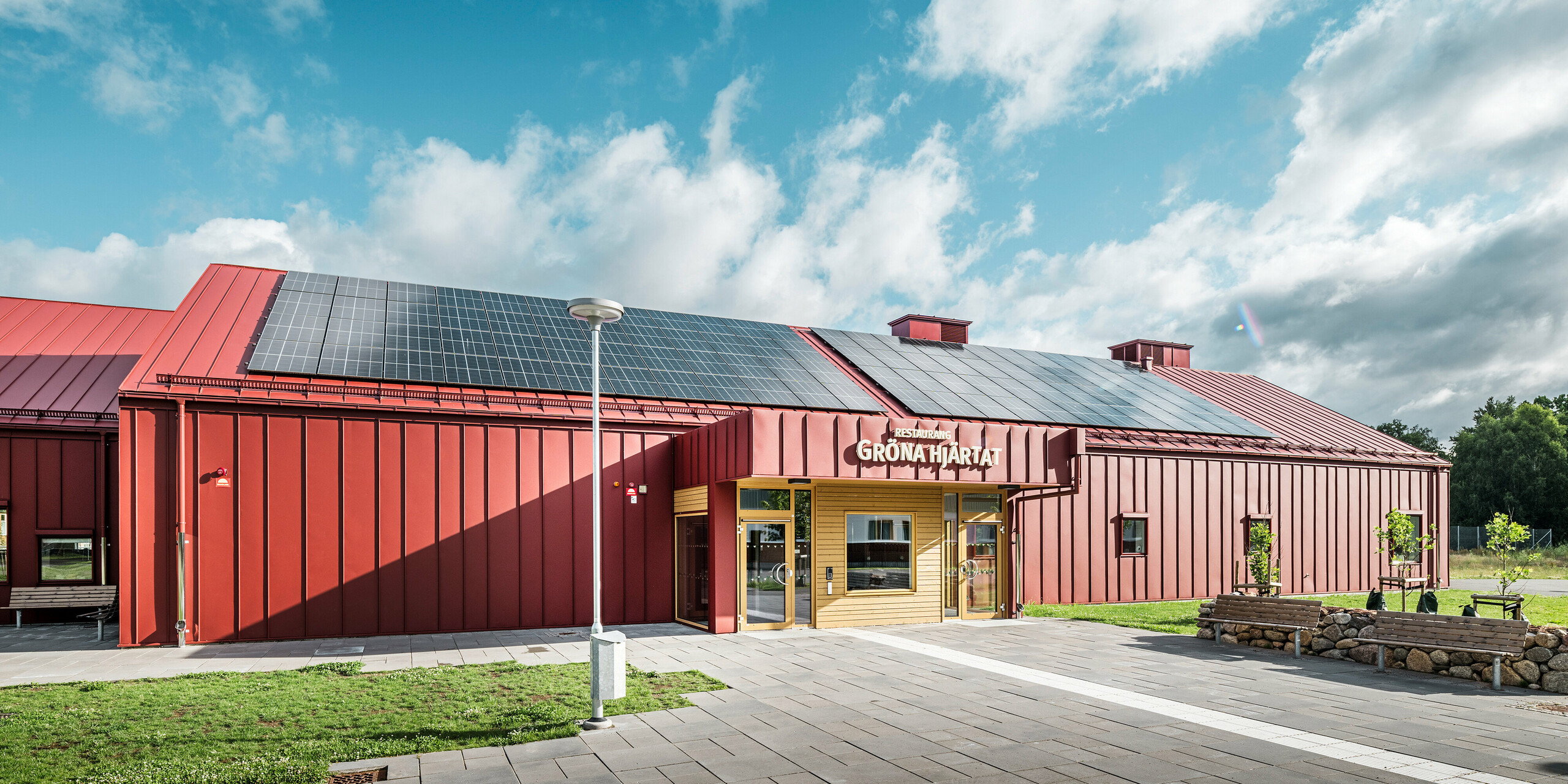 Blick auf den Eingangsbereich des Restaurants Gröna Hjärtat, Schulkantine der Göingeskolan und Prästavångskolan in Broby, Schweden. Das Gebäude wurde mit einer auffälligen Fassade aus PREFALZ in Oxydrot versehen. Auf dem Dach wurde eine großflächige Photovoltaikanlage installiert, die erneuerbare Energie liefert. Die Kombination aus langlebiger Aluminiumverkleidung und anspruchsvollem Design macht dieses Gebäude zu einem herausragenden Beispiel moderner Architektur. Die PREFA Dachentwässerung ist analog zu Dach und Fassade ebenfalls in Oxydrot ausgeführt und unterstreicht das harmonische Erscheinungsbild des BAuwerks.