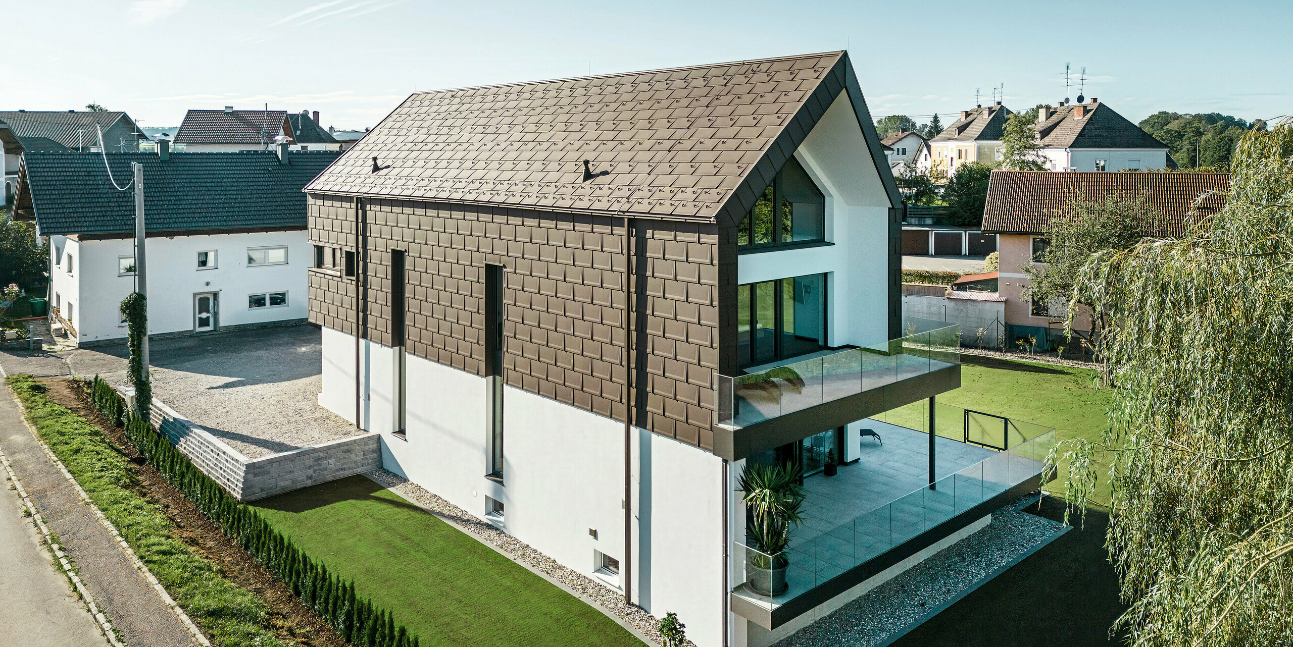 Luftaufnahme von der Straße eines modernen Einfamilienhauses in Neukirchen, Österreich. Das aktuelle Gebäude wurde an der Fassade großflächig mit PREFA R.16 Dachplatten in Dunkelbraun P.10 verkleidet. Die großen Fensterfronten und vertikalen Fenster, die sich fast über die gesamte Höhe des Hauses erstrecken, unterstreichen die einzigartige Architektur und wirken als Blickfang. Die außergewöhnliche Gebäudehülle wird durch eine schlichte weiße Putzfassade ergänzt. Um das Gebäude herum ist eine ruhige Wohngegend mit traditionellen Häusern zu sehen.