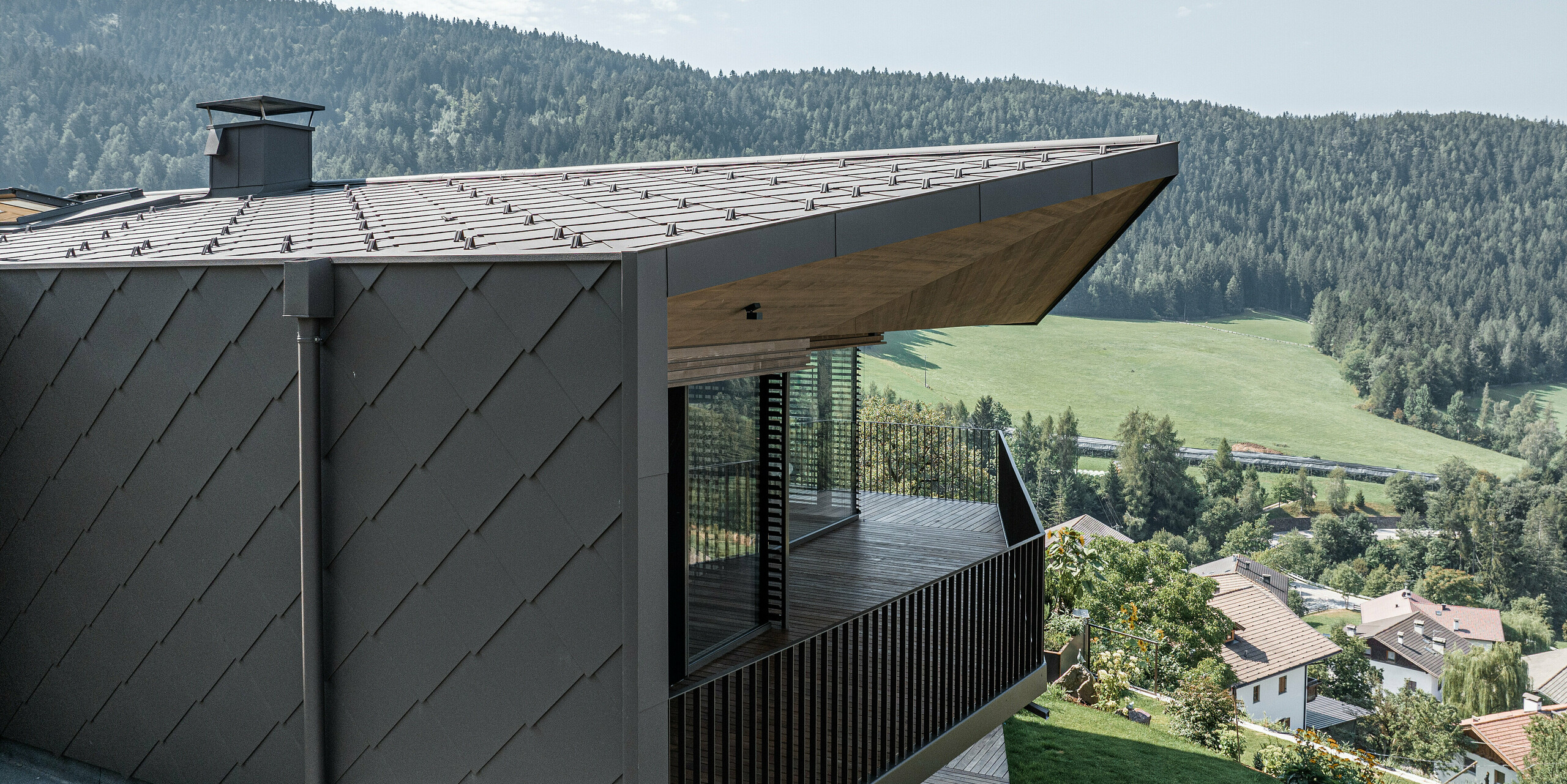Detailní záběr na roh modernizovaného dvojdomu v Möltenu v Jižním Tyrolsku s tmavě hnědými hliníkovými šablony od PREFA na střeše a fasádě. Návrh integruje velkou krytou terasu s dřevěným zábradlím, která nabízí úchvatné výhledy na okolní zelenou krajinu a vzdálené kopce. Pečlivě navržený přechod mezi materiály podtrhuje harmonické spojení přírody a moderní architektury. Budova byla původně postavena na počátku 90. V rámci rekonstrukce byla budova částečně přestavěna a částečně zrekonstruována.