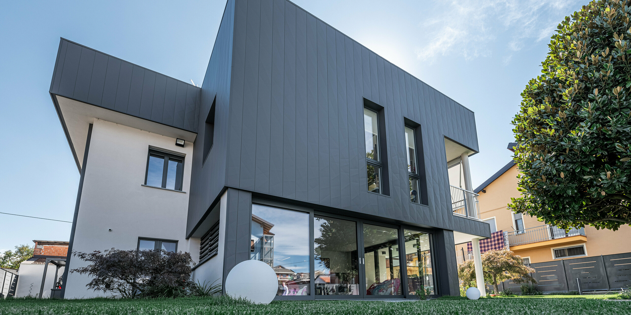 Perspektivische Ansicht eines modernen Einfamilienhauses in Savigliano, Italien, mit einer PREFA Siding.X Aluminiumfassade in P.10 Dunkelgrau. Das Foto, aufgenommen aus der Froschperspektive, zeigt die beeindruckende Architektur des Hauses, betont durch die klaren Linien und die dunkle, matte Fassade, die im Kontrast zum hellen Himmel und der üppigen grünen Umgebung steht. Große Fenster fördern die Integration des Innen- und Außenbereichs, wodurch ein lichtdurchfluteter Wohnraum geschaffen wird.