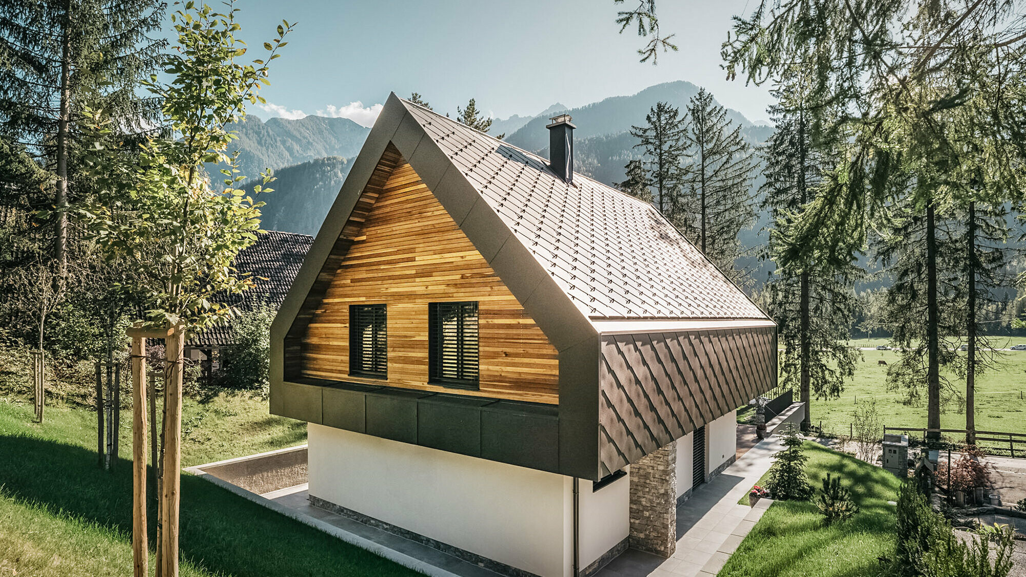 Ein modernes Chalet in Slowenien mit einer Kombination aus traditionellen und zeitgenössischen Architekturelementen, eingebettet in eine alpine Landschaft. Das Haus weist eine hölzerne Fassade mit geschlossenen Fensterläden auf, ein charakteristisches steiles Satteldach mit nussbraunen Dach- und Wandrauten und einen Schornstein. Das Anwesen ist umgeben von reichlich Grünflächen, Bäumen und bietet einen klaren Blick auf die entfernten Berge und ein offenes Tal