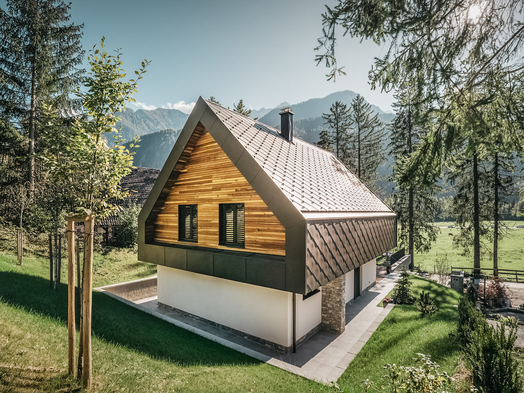 Ein modernes Chalet in Slowenien mit einer Kombination aus traditionellen und zeitgenössischen Architekturelementen, eingebettet in eine alpine Landschaft. Das Haus weist eine hölzerne Fassade mit geschlossenen Fensterläden auf, ein charakteristisches steiles Satteldach mit nussbraunen Dach- und Wandrauten und einen Schornstein. Das Anwesen ist umgeben von reichlich Grünflächen, Bäumen und bietet einen klaren Blick auf die entfernten Berge und ein offenes Tal