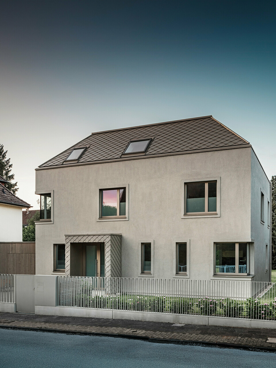 Ein modernes Einfamilienhaus mit Walmdach und bronze-farbener Aluminium-Dach- und Fassadenverkleidung von PREFA. Das Haus befindet sich in einer ruhigen Wohngegend, umgeben von gepflegten Gärten und anderen Einfamilienhäusern. Im Vordergrund ist ein niedriger Zaun zu sehen, der das Grundstück von der Straße trennt. Die Architektur des Hauses zeichnet sich durch klare Linien und große Fenster aus, die viel natürliches Licht in die Innenräume lassen. 