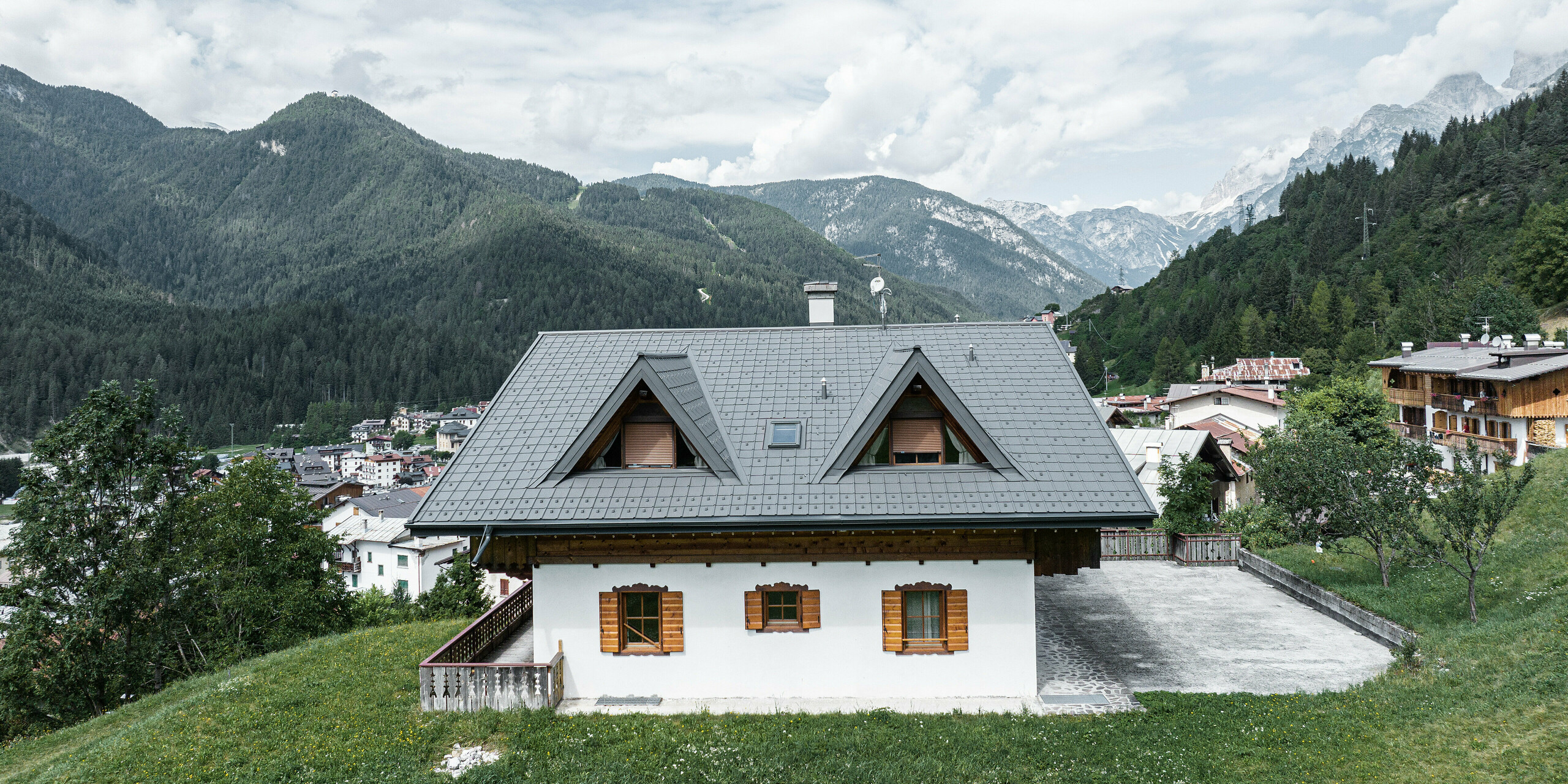 Die Rückseite einer Berghütte in Auronzo di Cadore, Italien, zeigt frisch verlegte PREFA Dachplatten in Hellgrau. Das Haus steht auf einer grünen Wiese mit den majestätischen Dolomiten im Hintergrund. Die beiden Spitzgauben fügen sich perfekt in die Bergkulisse im Hintergrund ein. Die sorgfältige Restaurierung des Daches unterstreicht den traditionellen Charme des Gebäudes, während moderne Elemente es mit der alpinen Landschaft verschmelzen.