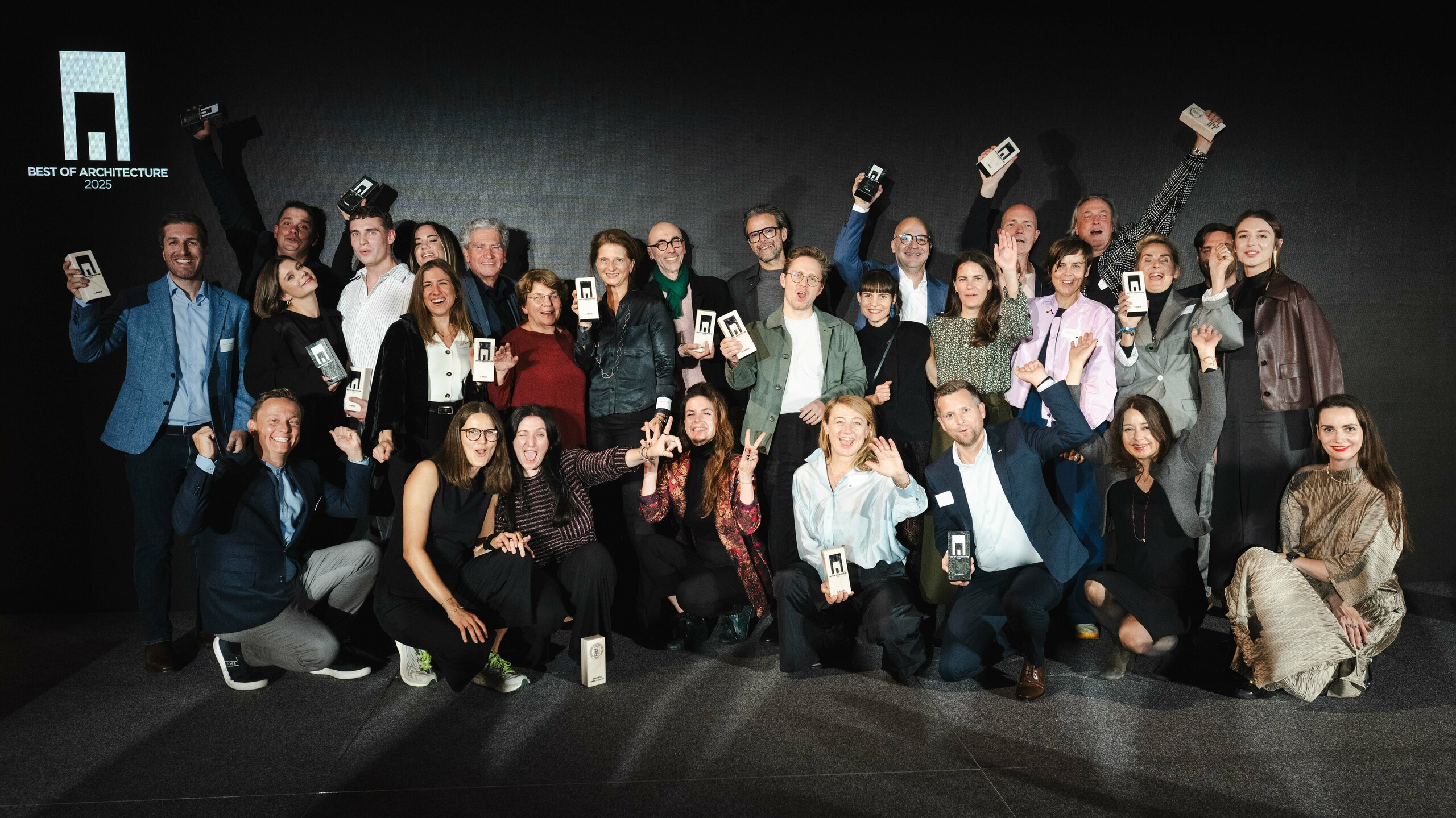 Gruppenfoto der Gewinner bei der Preisverleihung ‚Wohnbauten des Jahres‘ 2025: Die Preisträger stehen vor einer eleganten schwarzen Bühne mit dem Logo ‚Best of Architecture‘ und halten stolz ihre Auszeichnungen in den Händen. Dynamische Pose, moderne Kleidung und ein stimmungsvolles Lichtdesign unterstreichen die Bedeutung des Events. Die Szene symbolisiert Innovation und Architekturqualität.