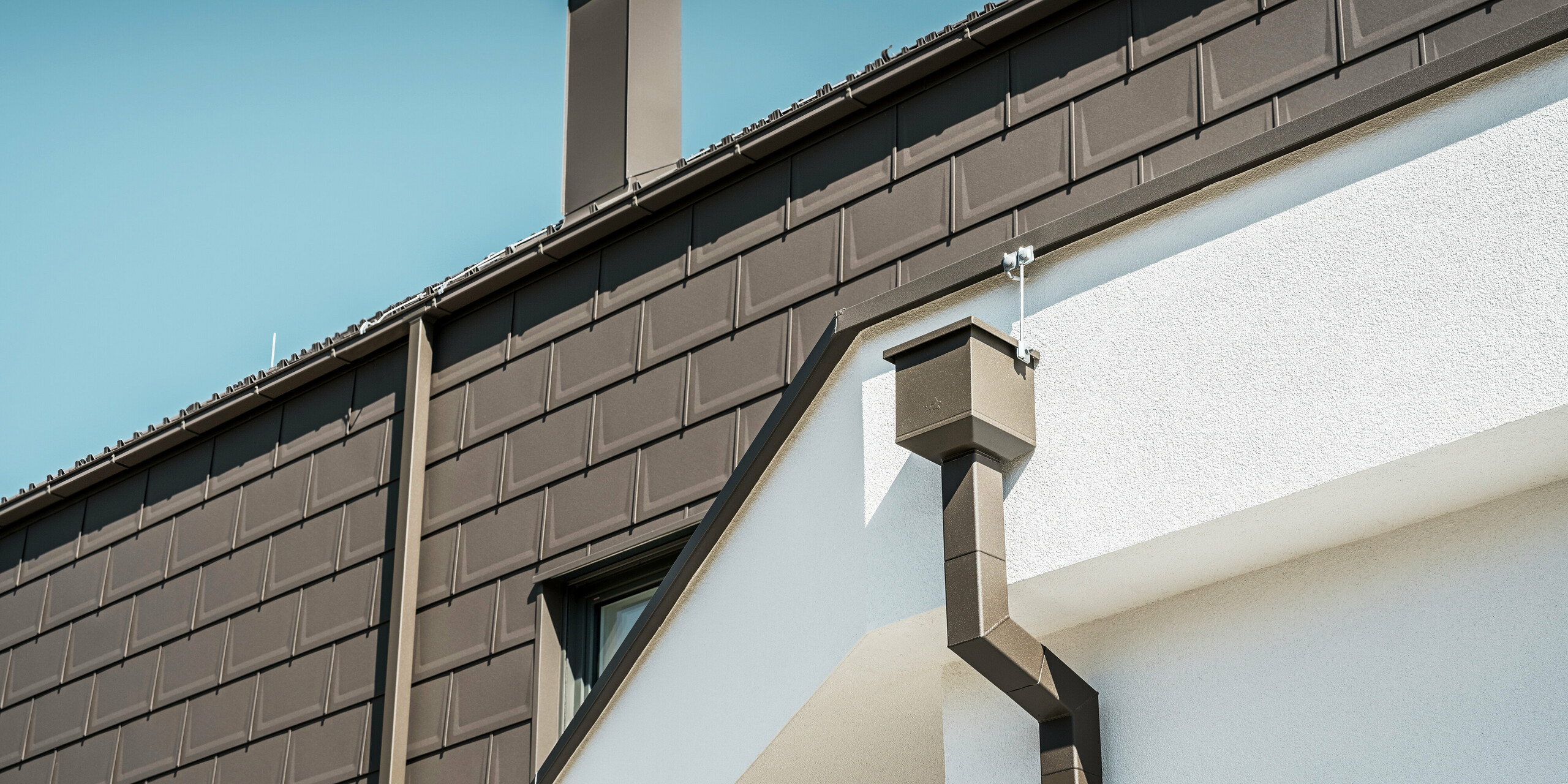 Detailaufnahme des Dachrandes bzw. der Fassade eines Hauses in Neukirchen, Österreich, ausgestattet mit PREFA R.16 Dachplatten in P.10 braun. Sowohl die Dachplatten, die bei diesem Gebäude als Dach- und Fassadenverkleidung dienen, als auch die Dachentwässerung, bestehend aus Vierkantrohr, Wassersammelkasten und Kastenrinne, sind in der gleichen Farbe gehalten und bieten eine einheitliche, hochwertige Ausführung. Die Verwendung von PREFA Produkten verleiht dem Gebäude ein zeitgemäßes Aussehen, während die ästhetische Einbindung der Entwässerungselemente die funktionale Eleganz des Designs unterstreicht.