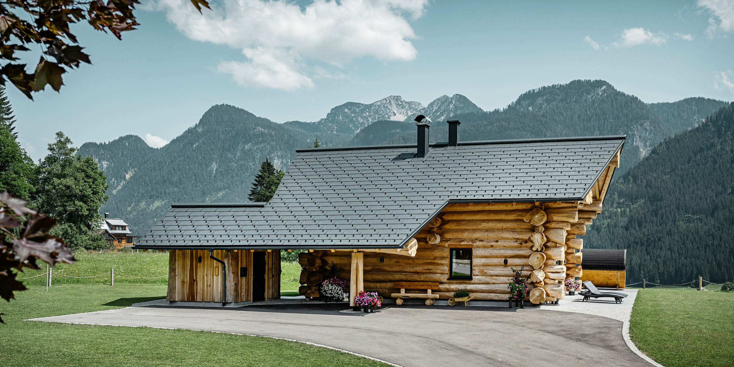 Blockhaus in Gosau, Österreich, mit einem PREFA Dachsystem in der Farbe Anthrazit. Die bewährten Dachplatten R.16 schaffen einen ansprechenden Kontrast zum natürlichen Holz des Blockhauses. Im Hintergrund erstreckt sich ein majestätisches Bergpanorama, das die ruhige und malerische Lage des Hauses unterstreicht. Die detailreiche Aufnahme fängt die Symbiose aus traditioneller Baukunst aus Österreich und moderner Dachtechnologie ein und zeigt die robuste und ästhetische Qualität der PREFA Aluminiumprodukte.