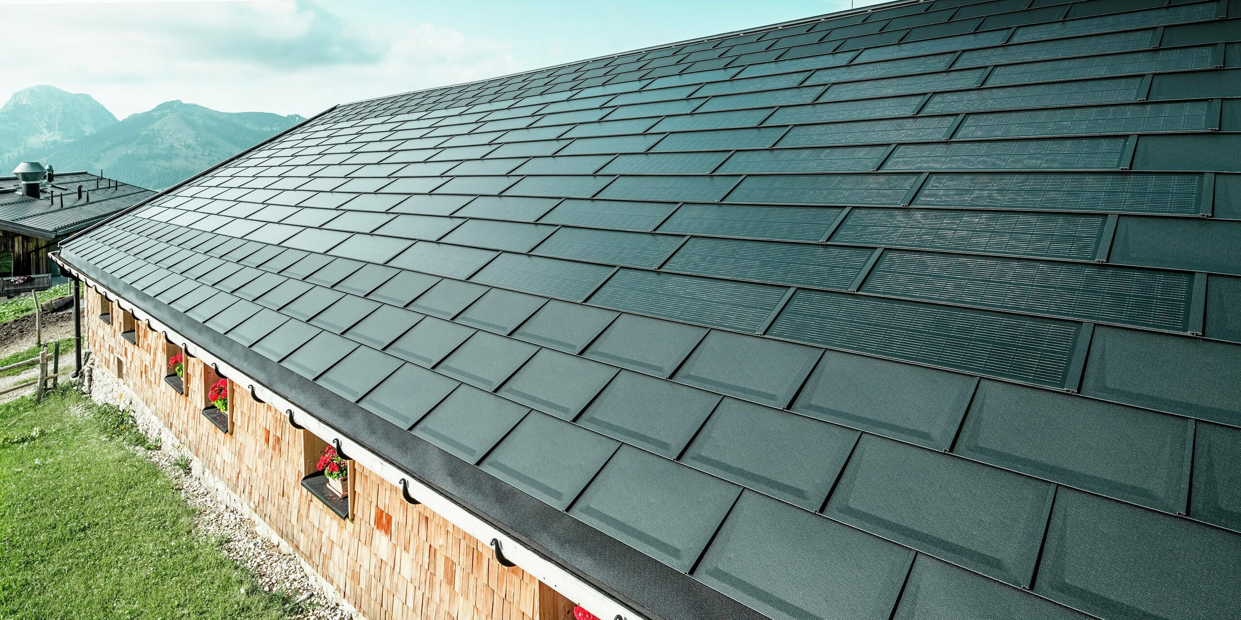 Nahaufnahme der Birkehütte auf der Walleralm in Bayrischzell mit neuem Blechdach aus hochwertigen PREFA Dachplatten R.16 in P.10 Schwarz und modernen PREFA Solardachplatten. Die Dachsanierung kombiniert eine zeitgemäße Photovoltaik-Anlage mit einer widerstandsfähigen Aluminium-Dacheindeckung, die optimalen Schutz vor alpinen Witterungseinflüssen bietet. Die traditionellen Holzschindeln an der Fassade und die Blumenfenster verleihen der Hütte einen charmanten, rustikalen Charakter.