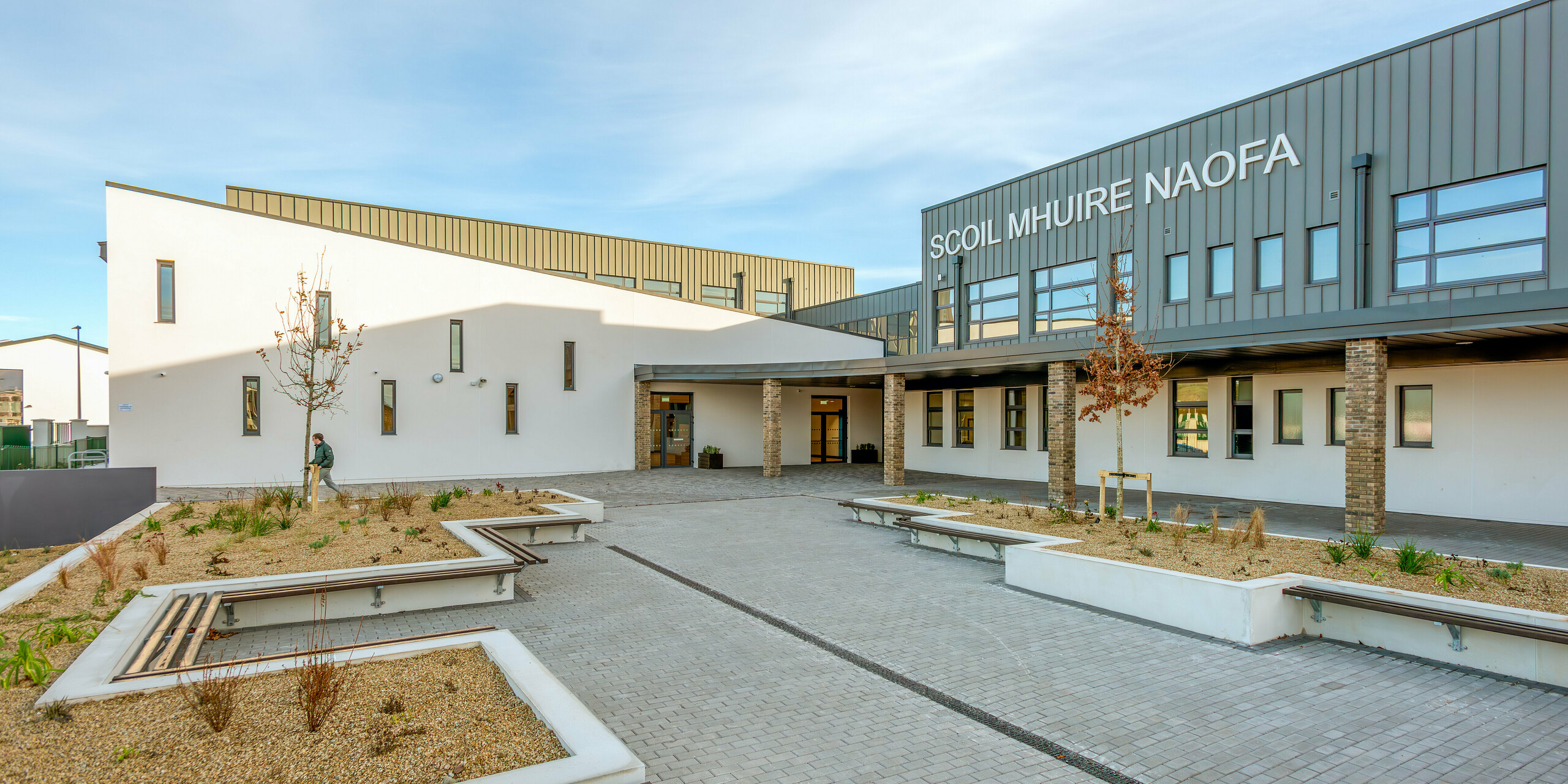 Einladender Außenbereich der Scoil Mhuire Naofa in Carrigtwohill, Irland, mit Sitzgelegenheiten und gepflegter Bepflanzung. Die Fassade aus PREFALZ Aluminium in P.10 Hellgrau verleiht dem Schulgebäude eine langlebige und witterungsbeständige Eleganz, die durch klare Linien und funktionales Design ergänzt wird.