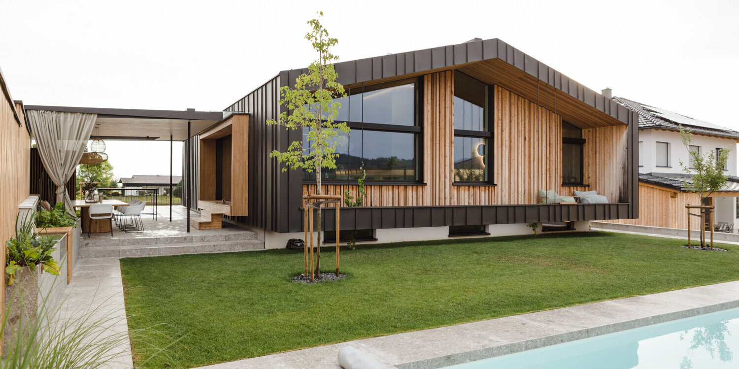 Idyllisches Einfamilienhaus in Kirchham, Österreich, mit einer Fassade aus PREFALZ in P.10 Anthrazit, die nahtlos in die umgebende Garten-Landschaft übergeht. Das Holz und Glas bieten eine natürliche Wärme und Transparenz, während die klaren Linien des Aluminiums von PREFA eine moderne Eleganz ausstrahlen. Der gepflegte Garten mit einem einladenden Poolbereich ergänzt das Wohlfühlambiente des Hauses und macht es zu einem perfekten Rückzugsort für zeitgenössisches Wohnen.