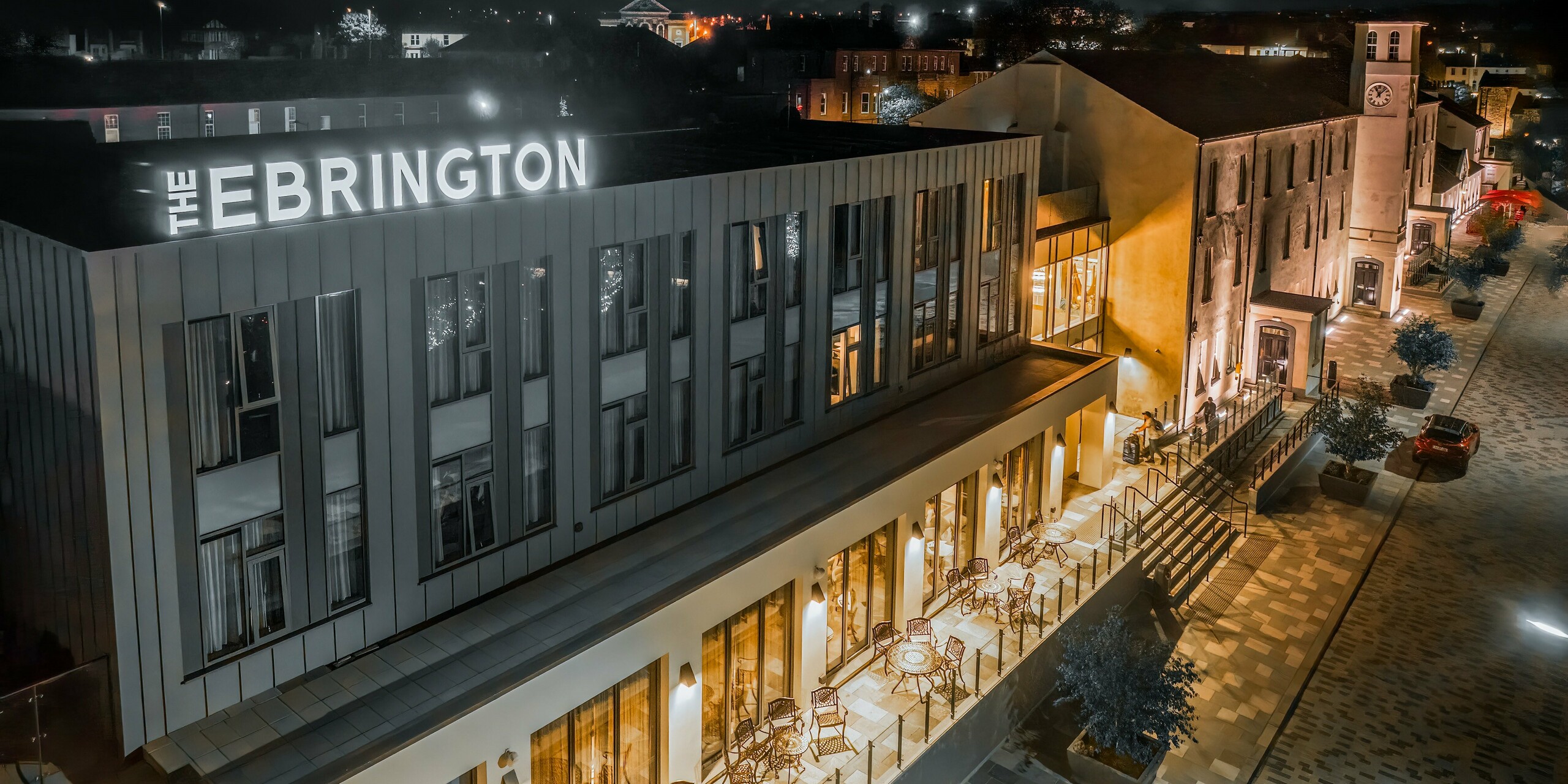 Das Ebrington Hotel bei Nacht besticht durch seine eindrucksvolle Beleuchtung in einer Fußgängerzone im Zentrum von Londonderry, Nordirland.  Das moderne Gästehaus wurde mit einer robusten PREFALZ Verkleidung aus Aluminium in den Farben P.10 Lichtgrau und P.10 Zinkgrau verkleidet. Diese hochwertige Blechfassade schützt das Gebäude vor Witterungseinflüssen. Zudem wirkt die Stehfalzdeckung als Highlight in der schlichten Architektur.