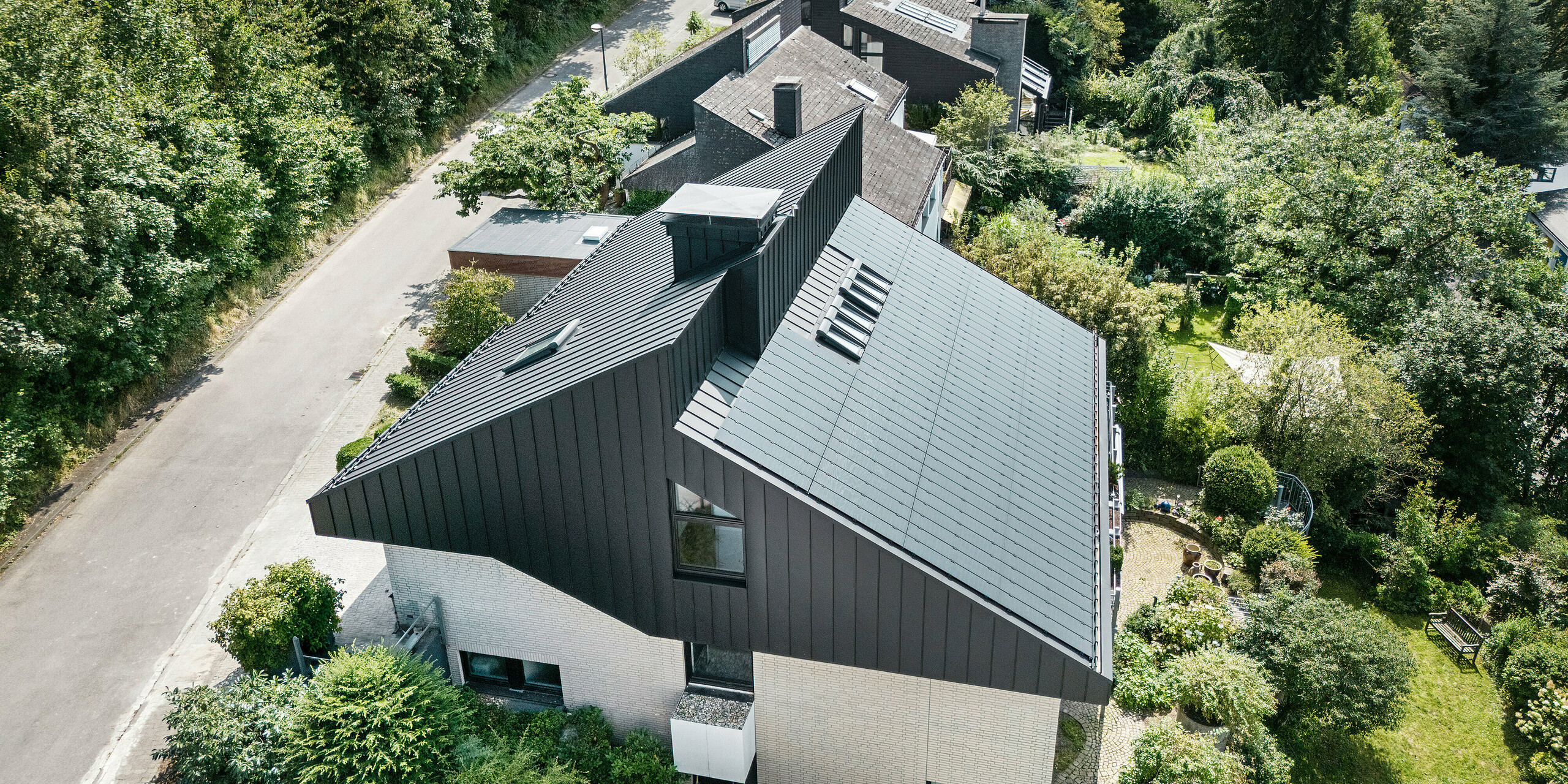 Schrägansicht auf ein modernes Einfamilienhaus in Meschede, Deutschland. Das versetzte Pultdach wurde mit PREFALZ in P.10 Schwarz verkleidet. Das Aluminiumdach dient als optisches Highlight und bietet dem Gebäude dauerhaften Schutz. Die präzise Doppelstehfalzdeckung zieht sich vom Dach über die Fassade an den Giebelseiten. Umgeben von üppigem Grün und einer ruhigen Straße fügt sich das energieeffiziente Gebäude mit den innovativen PREFALZ Solarmodulen perfekt in das naturnahe Wohnumfeld ein.