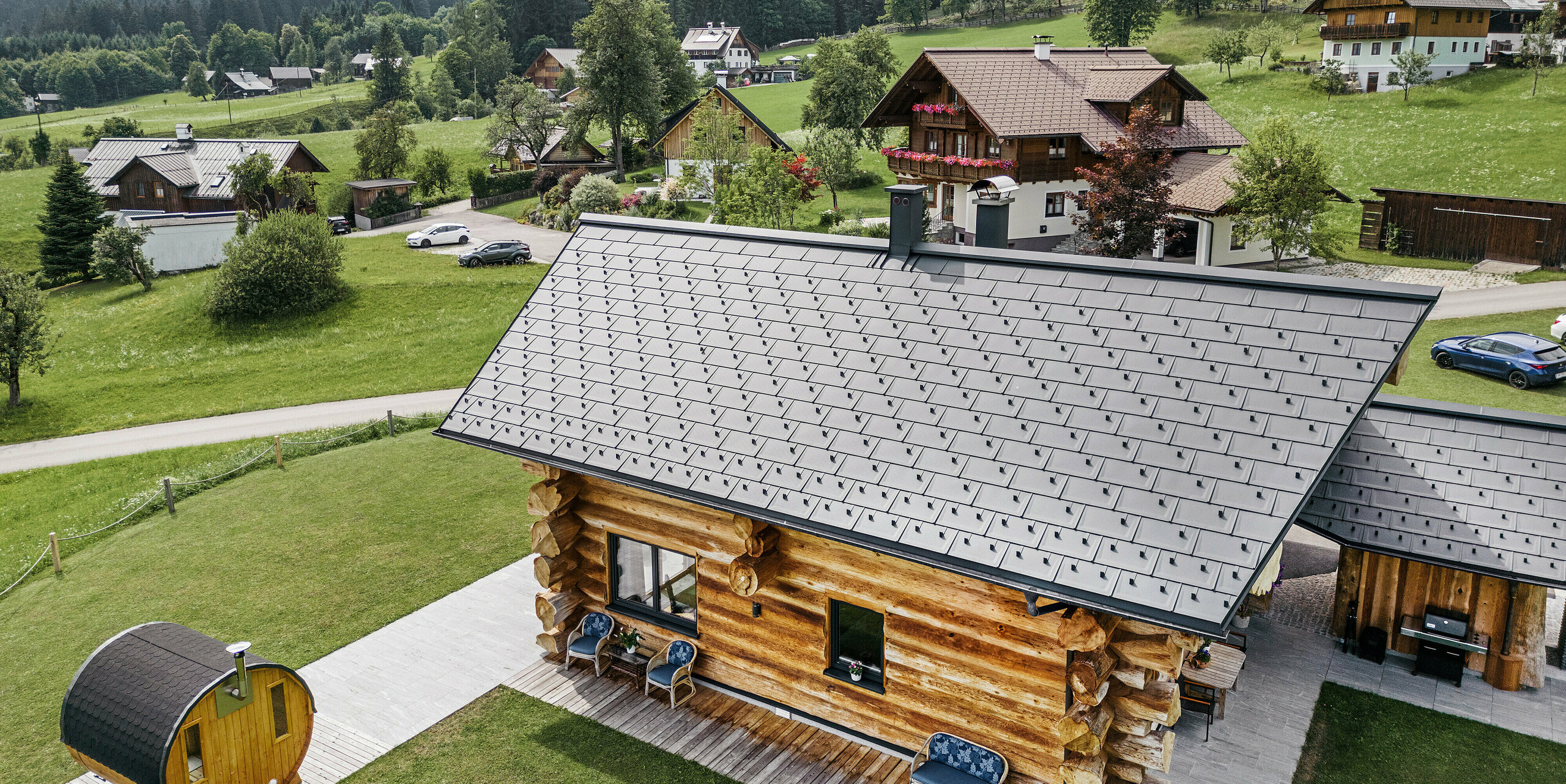 Vogelperspektive eines rustikalen Blockhauses in Gosau, Österreich, mit einem hochwertigen PREFA Aluminiumdach aus Dachplatten R.16 in P.10 Anthrazit. Das Dach zeigt die typische Struktur und Präzision der PREFA Dachsysteme, die sich optimal in die ländliche Umgebung einfügen. Das satte Grün der Wiesen, die umliegenden traditionellen Häuser und die bewaldeten Hügel im Hintergrund bilden einen natürlichen Kontrast zu den modernen, anthrazitfarbenen Dachelementen, die Beständigkeit und moderne Ästhetik in die alpine Architektur bringen.