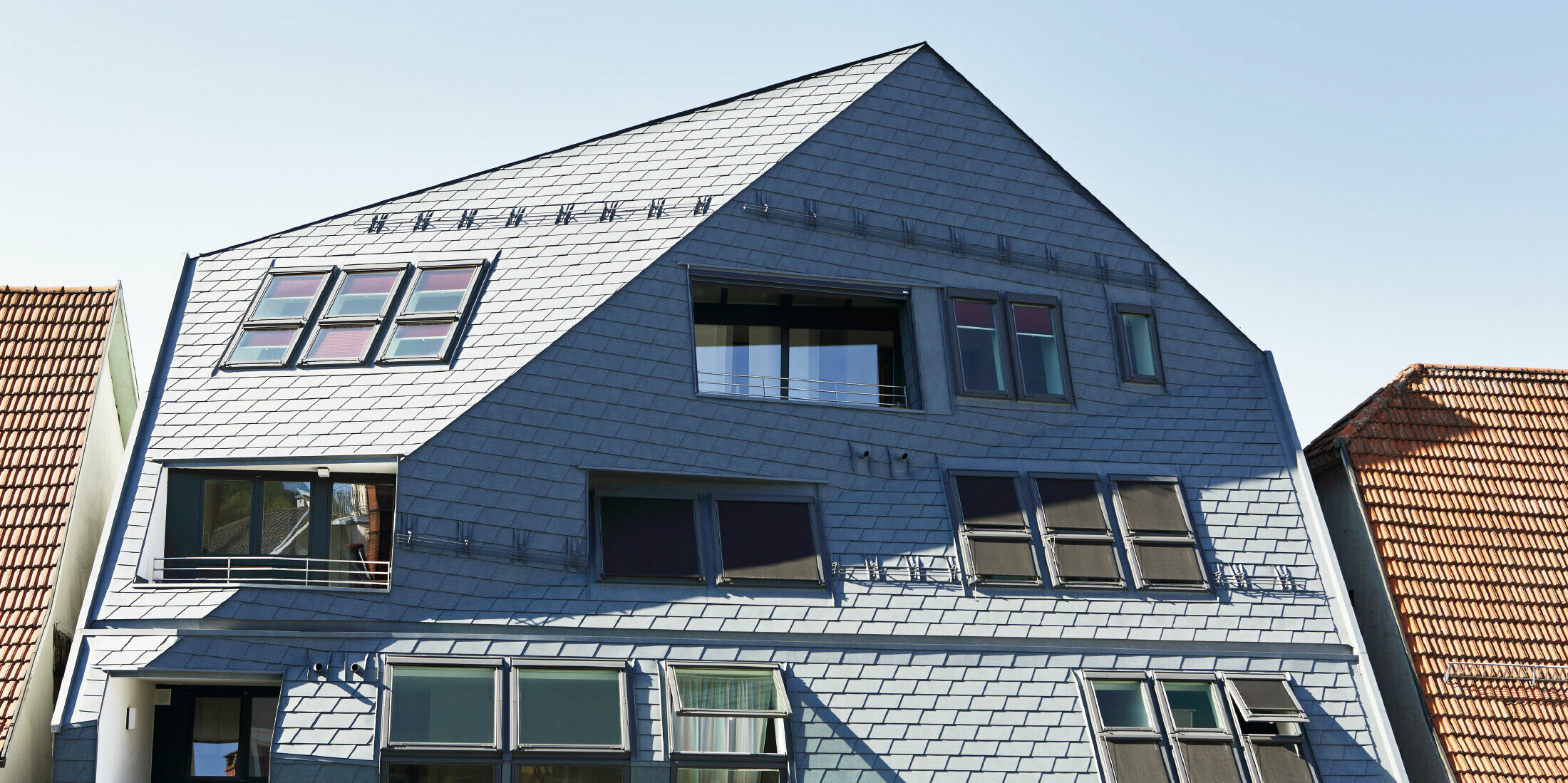 Galeriehaus mit gekanteter Fassadenfläche, verkleidet mit der PREFA Dach- und Wandschindel in P.10 Steingrau mit vielen Fenstern