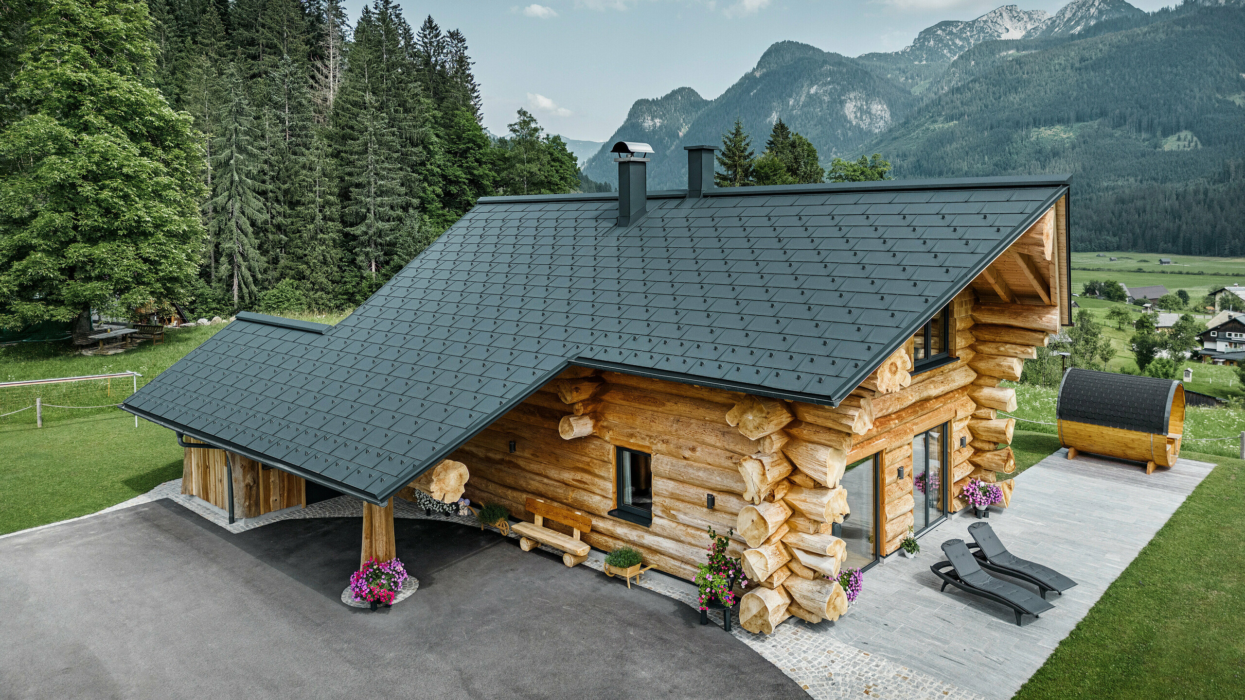 Schräge Ansicht eines malerischen Blockhauses in Gosau, Österreich, ausgestattet mit einem PREFA Dach aus anthrazitfarbenen Dachplatten R.16. Die Architektur kombiniert rustikales Blockhausflair mit modernen Elementen, umgeben von dichten grünen Wäldern und einer beeindruckenden Bergkulisse. Das Haus strahlt mit seiner ländlichen Eleganz und den präzise verlegten Dachelementen aus Aluminium, die für ihre Langlebigkeit und Witterungsbeständigkeit bekannt sind, eine einladende Atmosphäre aus.