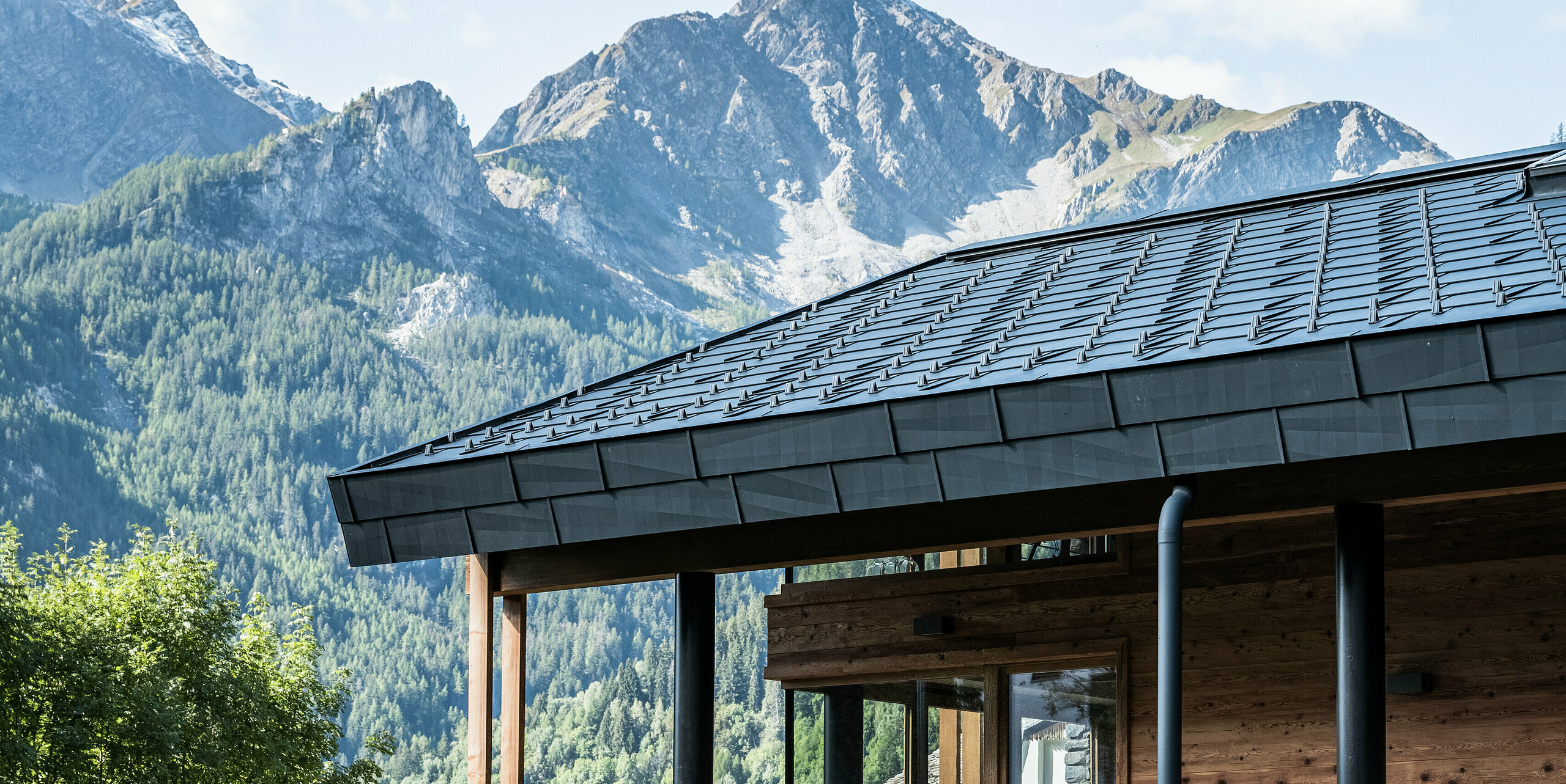 Eine Detailaufnahme des Daches einer modernen Villa in Courmayeur mit einer Fassade aus Holz, Stein und Blech. PREFA FX.12 Dachplatten in P.10 Steingrau ergänzen die strukturierte Architektur vor der malerischen Berglandschaft.