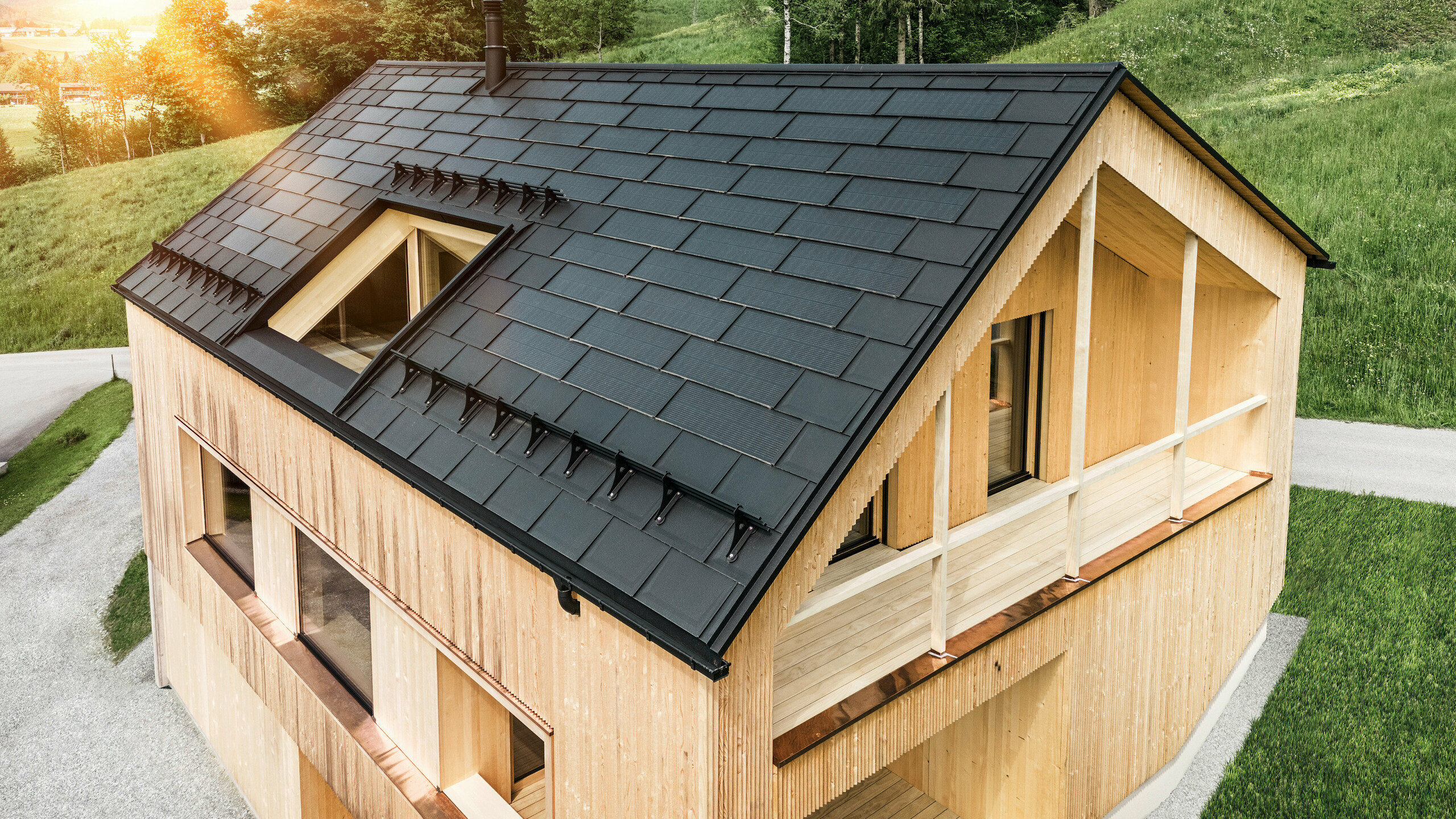 Blick auf ein modernes, nachhaltiges Holzhaus in Egg mit einem PREFA Solardach, das durch seine dunkle, strukturierte sowie elegante Erscheinung auffällt. Die Solarmodule sind strategisch zwischen den klassischen Dachplatten R.16 platziert, um eine optimale Sonneneinstrahlung zu nutzen und sich dabei ästhetisch in das Gesamtdesign einzufügen. Das Haus steht im Einklang mit seiner grünen Umgebung, während die warme Abendsonne die natürliche Holztextur der Fassade unterstreicht und die solide Bauweise betont. Ein harmonisches Beispiel für umweltbewusstes Bauen und Energieeffizienz.