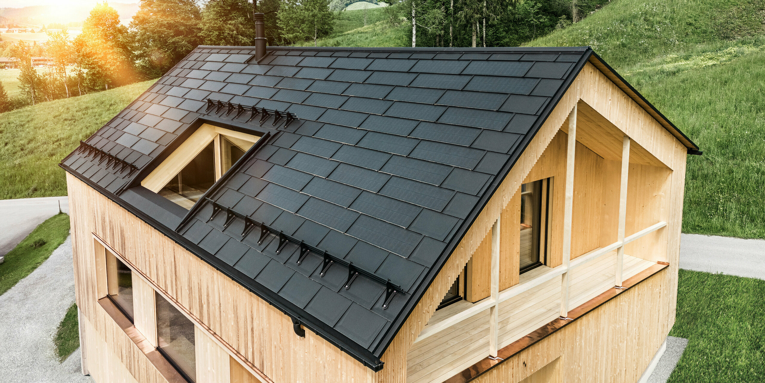 Blick auf ein modernes, nachhaltiges Holzhaus in Egg mit einem PREFA Solardach, das durch seine dunkle, strukturierte sowie elegante Erscheinung auffällt. Die Solarmodule sind strategisch zwischen den klassischen Dachplatten R.16 platziert, um eine optimale Sonneneinstrahlung zu nutzen und sich dabei ästhetisch in das Gesamtdesign einzufügen. Das Haus steht im Einklang mit seiner grünen Umgebung, während die warme Abendsonne die natürliche Holztextur der Fassade unterstreicht und die solide Bauweise betont. Ein harmonisches Beispiel für umweltbewusstes Bauen und Energieeffizienz.