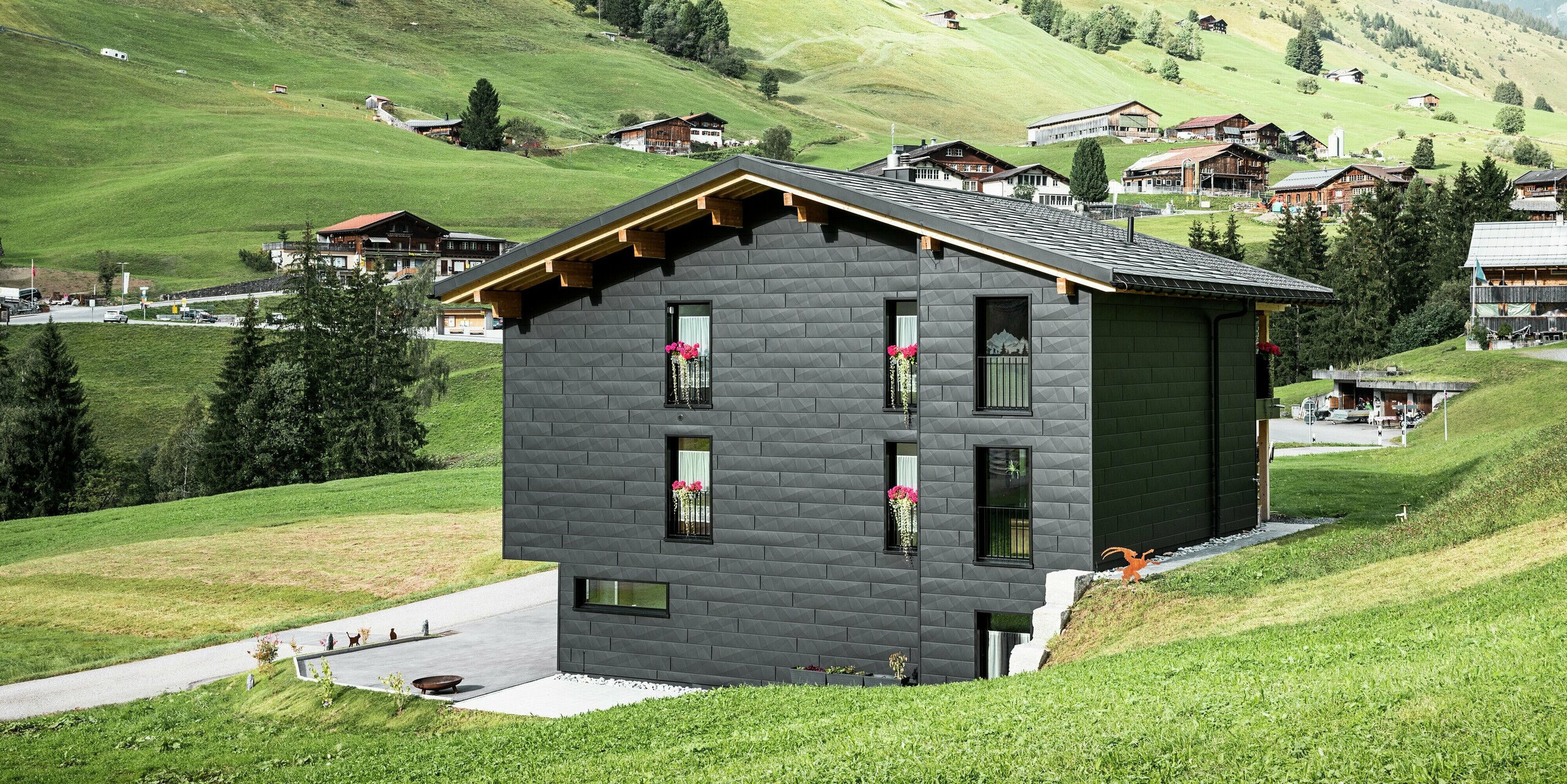 Rückansicht des Einfamilienhauses in Ascharina, Schweiz, mit einer markanten Aluminiumfassade aus PREFA Siding.X in P.10 Schwarz. Die Längs- und Querkantungen der Aluminiumpaneele verleihen dem Gebäude eine einzigartige und moderne Struktur. Die großen Fenster fangen viel Licht ein und harmonieren mit den natürlichen Holzelementen, die die Fassade akzentuieren.